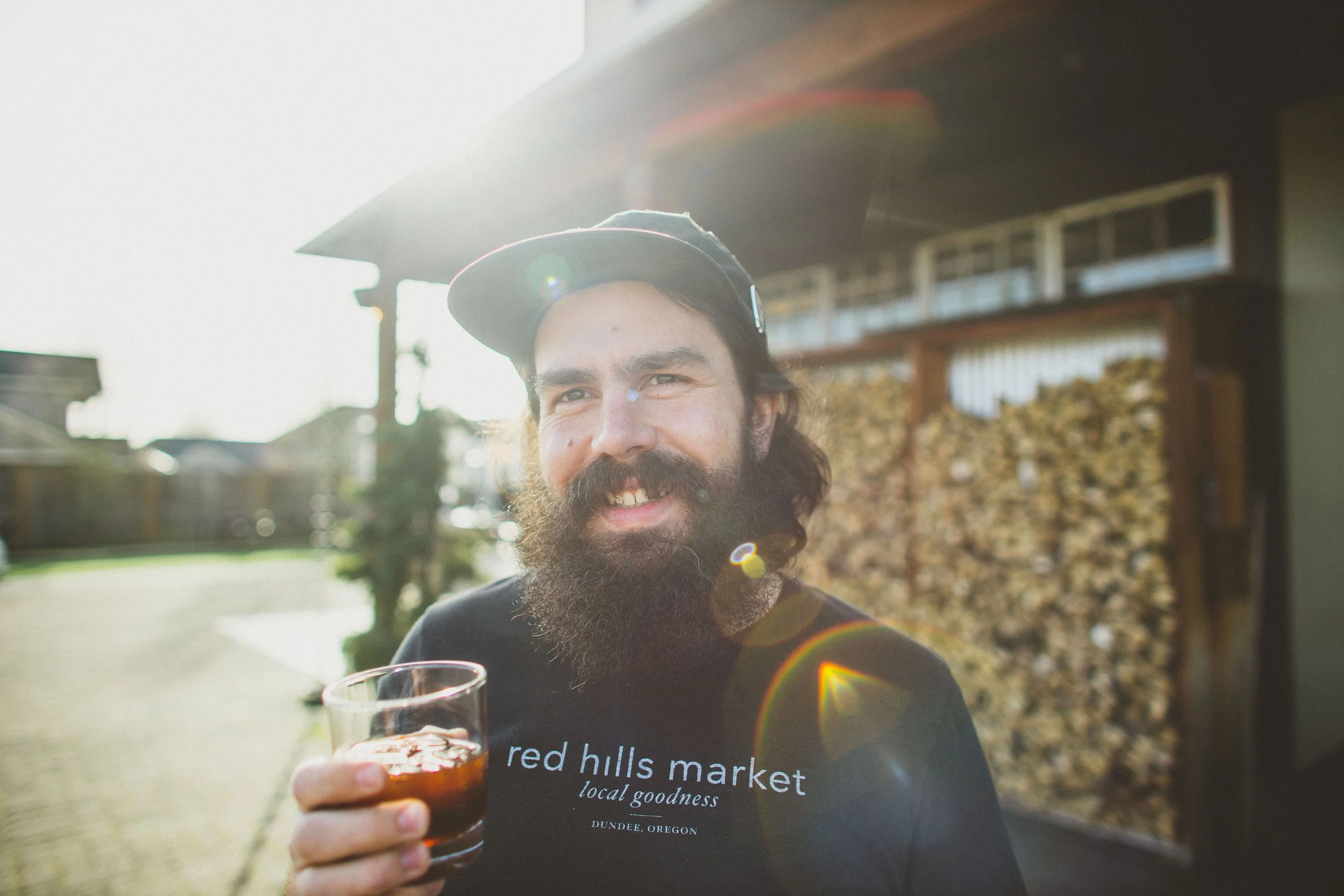 A man with a beard and mustache wearing a black cap and a black shirt that reads 'red hills market, local goodness, Dundee, Oregon' is holding a glass of dark beverage outdoors, with sunlight and a slightly blurred background of a wooden structure and a stack of wood.