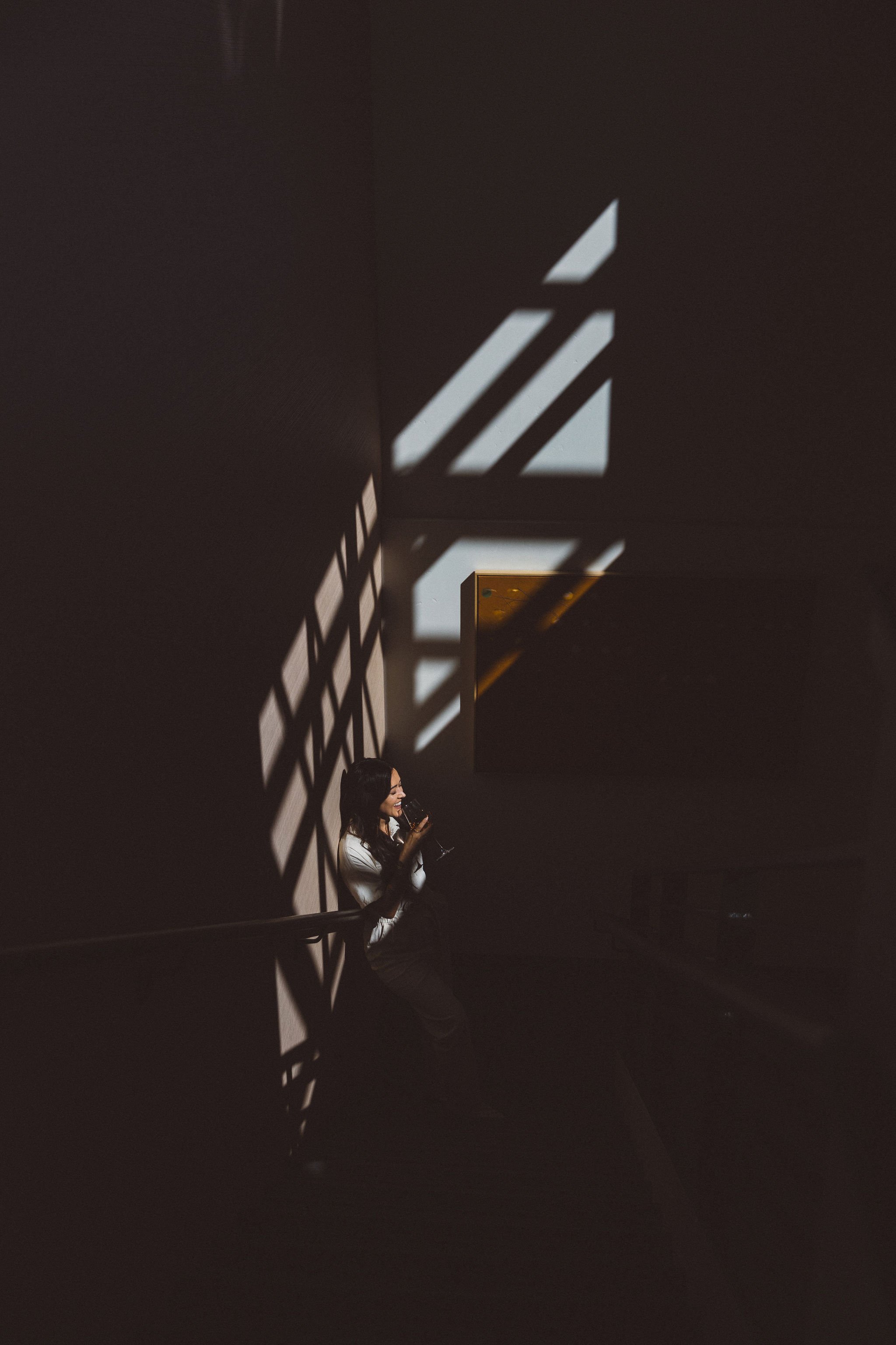A woman business owner sitting on a stairway in a dark room in Boise Idaho during a branding photo session, drinking from a glass with shadow patterns shaped by sunlight streaming through a window.