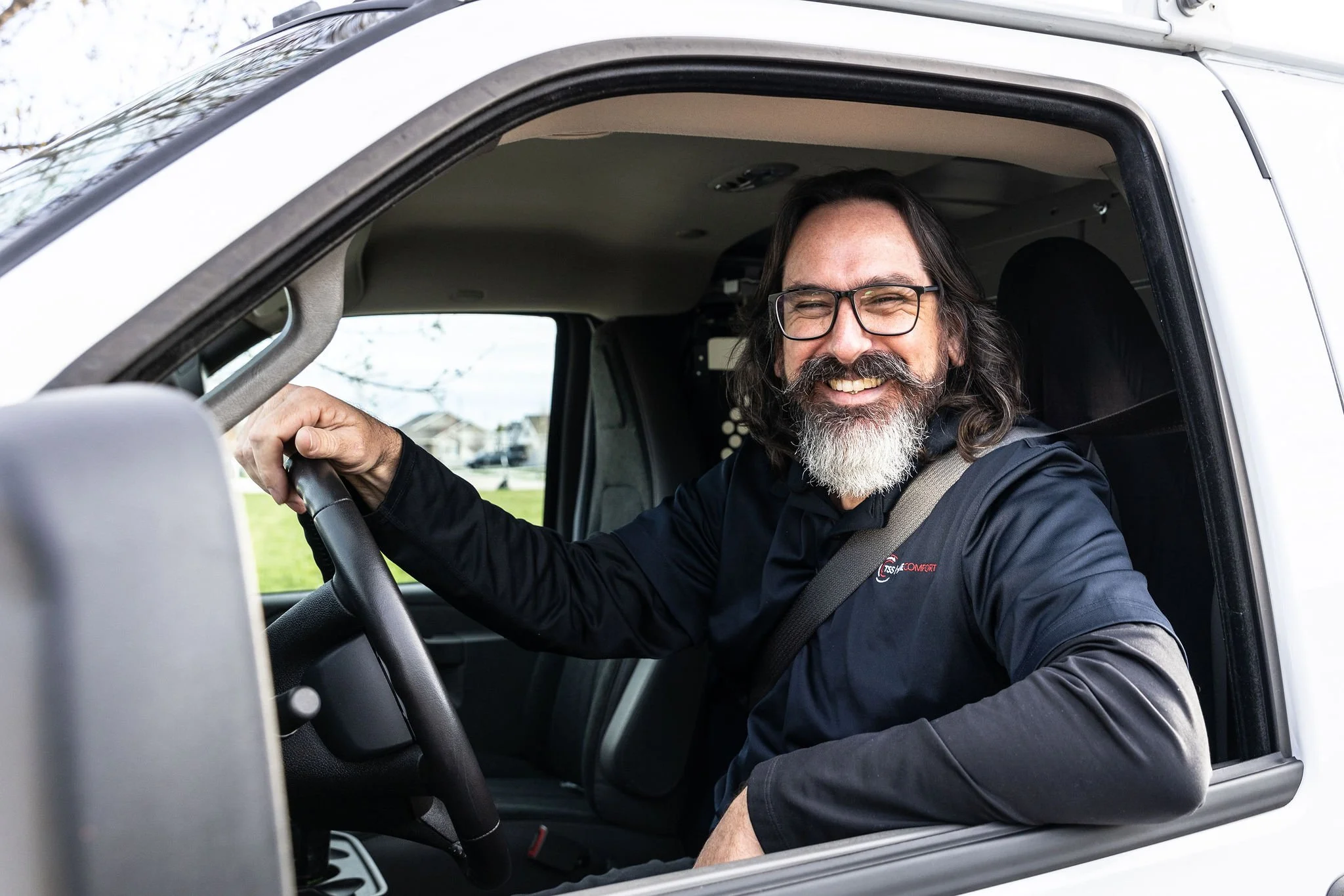 A smiling middle-aged man with glasses and a beard sitting in the driver's seat of a vehicle, looking out the window.