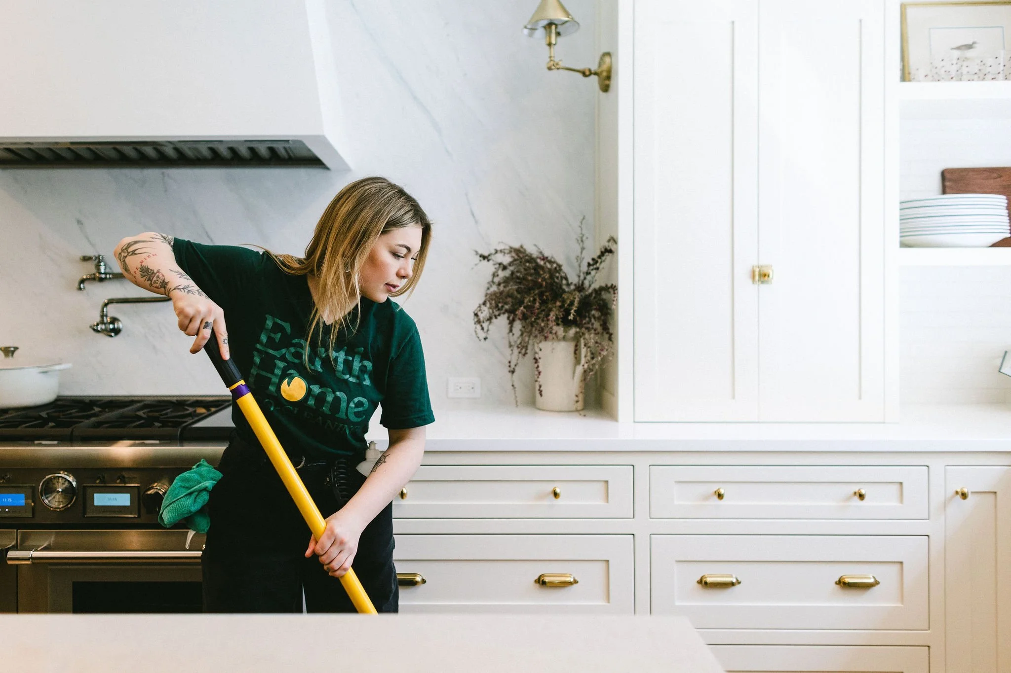 Woman with tattoos cleaning a kitchen counter with a yellow mop, wearing a green T-shirt with text, in a white kitchen with gold hardware and a flower vase in the background.