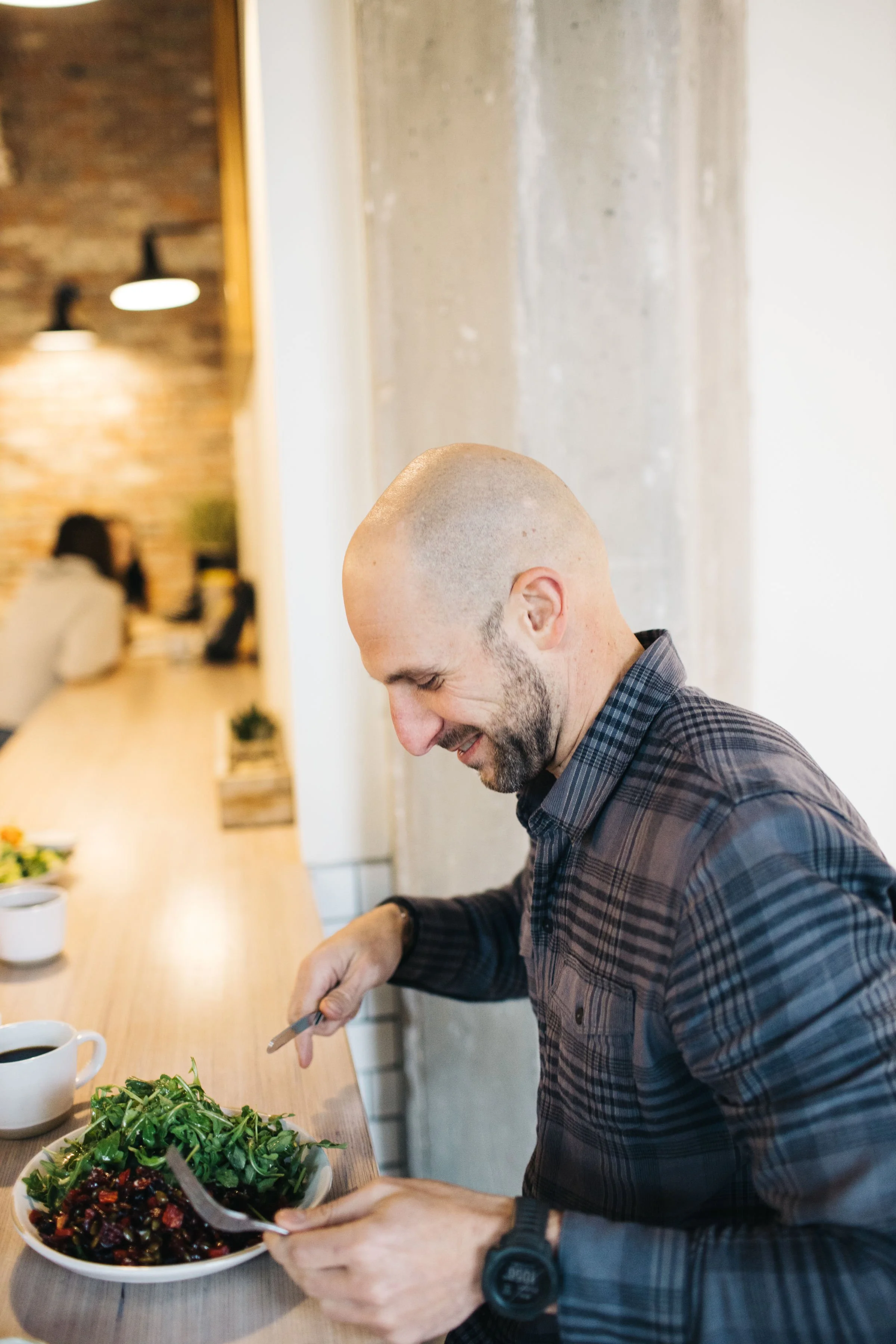 A man with a beard and shaved head smiling as he serves himself a salad at a restaurant or cafe during a branding photoshoot in downtown Boise.