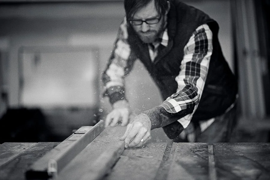Black and white creative lifestyle portrait of a carpenter cutting a board on a table saw in a workshop during a personal branding photoshoot in Boise, Idaho.