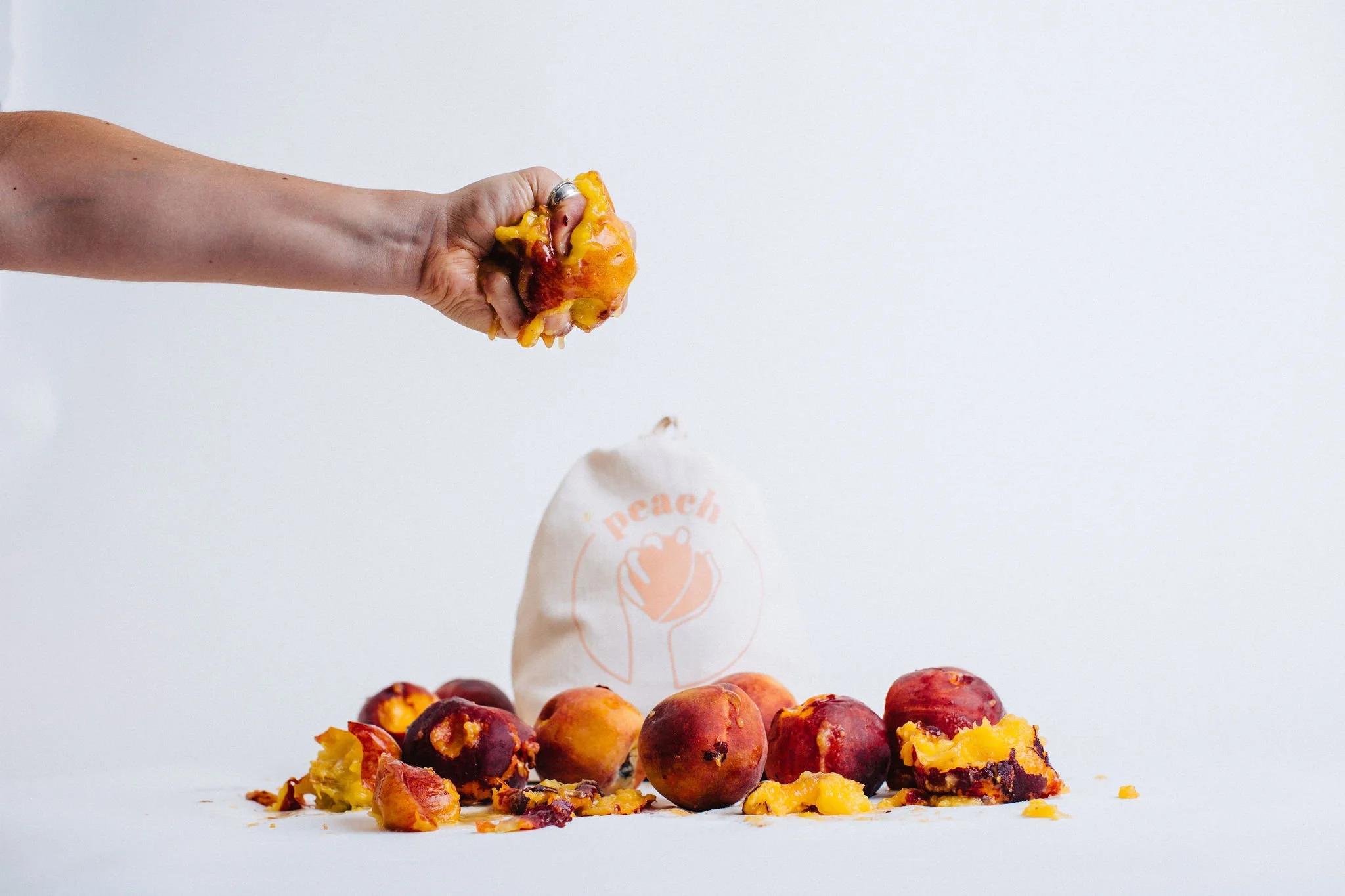 A person's hand squeezing a peach over a pile of spoiled peaches, with some spilling onto a white surface and a small cloth bag with an orange peach logo in the background during a branding studio photoshoot in Boise Idaho.