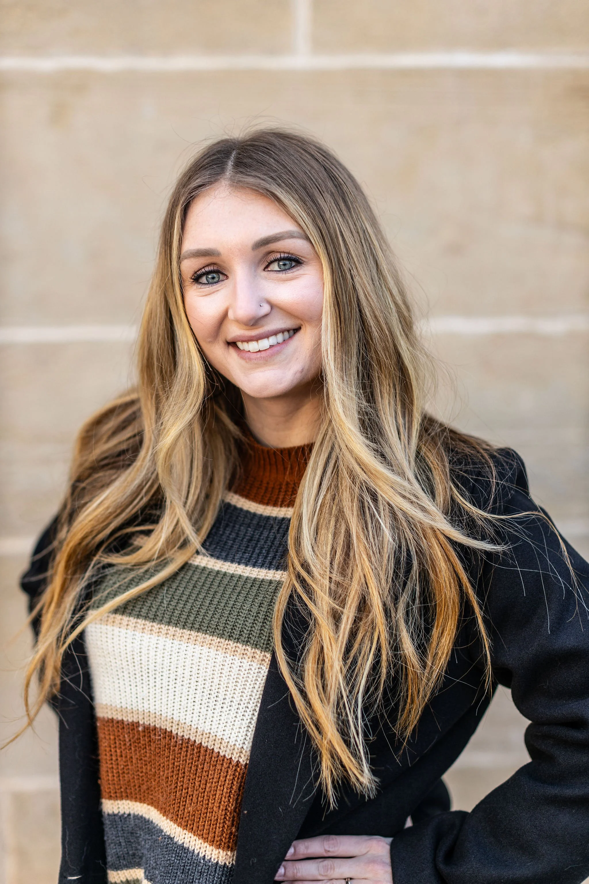 A smiling woman with long, wavy blonde hair, wearing a patterned sweater and a black coat, posing outdoors in front of a beige stone wall.