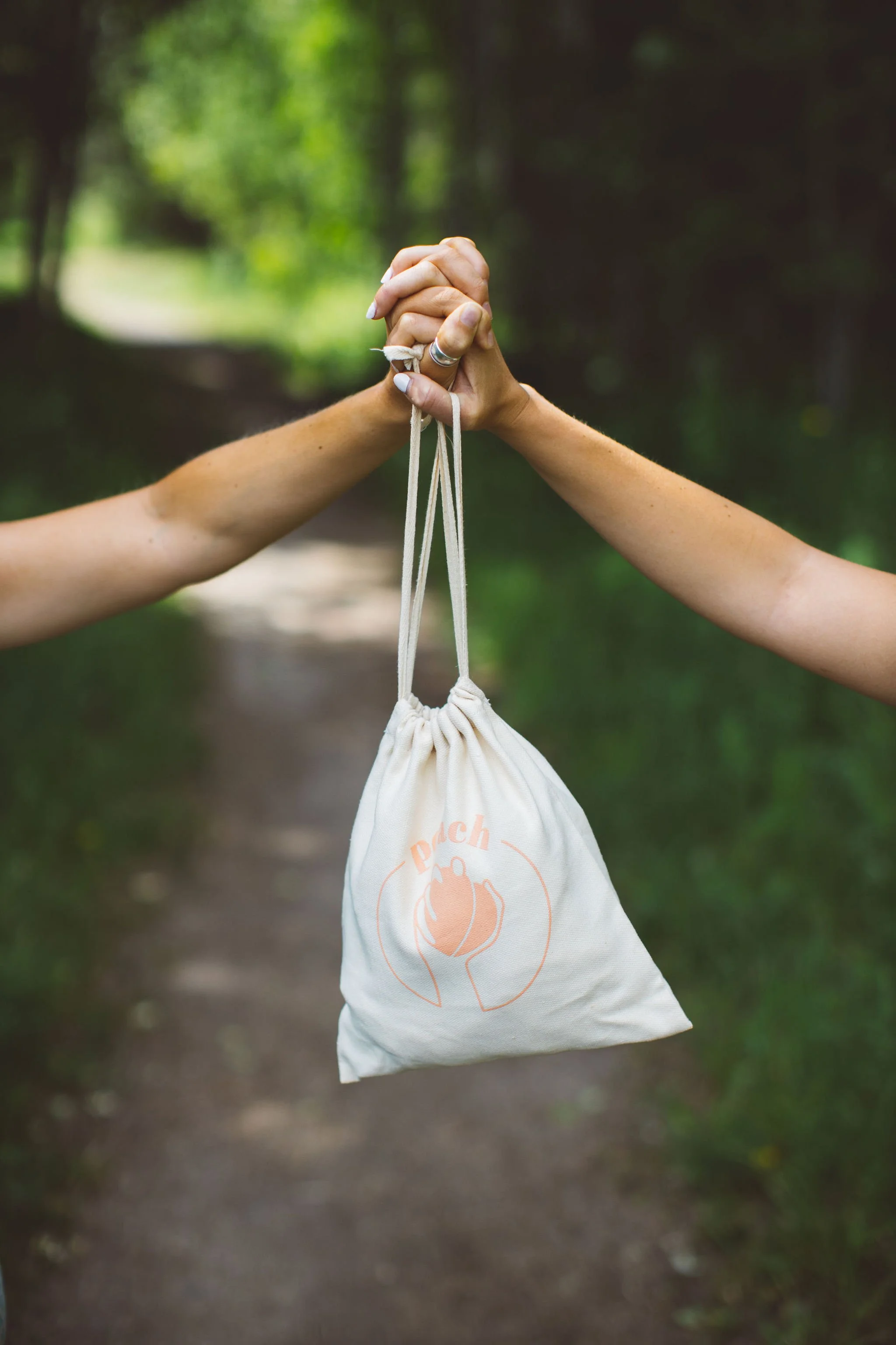 Two hands clasped together hold a small cloth bag with a peach logo and the word 'peach' on it, in a forest setting during a branding photoshoot in Boise, Idaho.