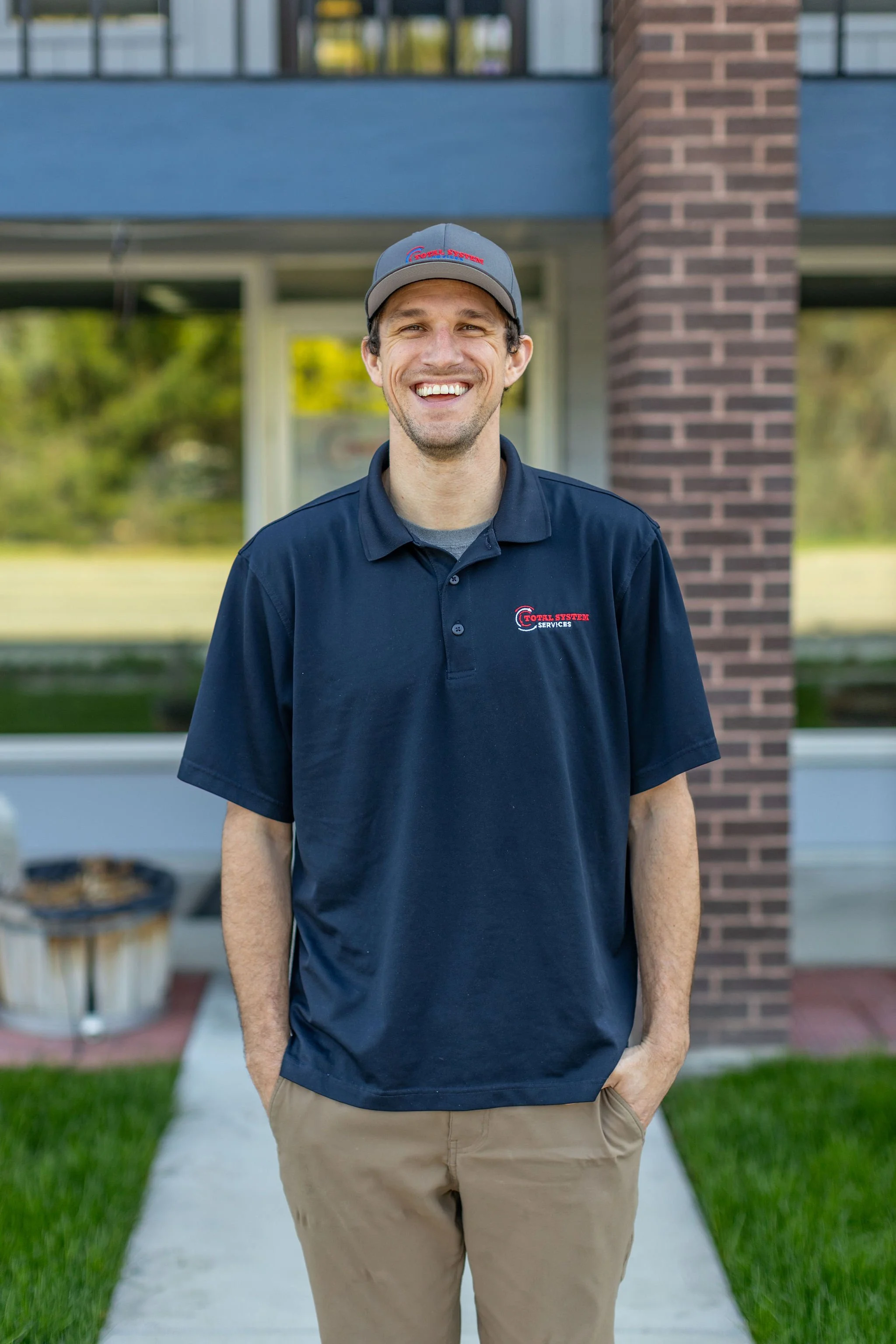A smiling man, wearing a navy polo shirt and a gray cap, stands outside in front of a building with a brick pillar and windows, with greenery and a sidewalk visible in the background.