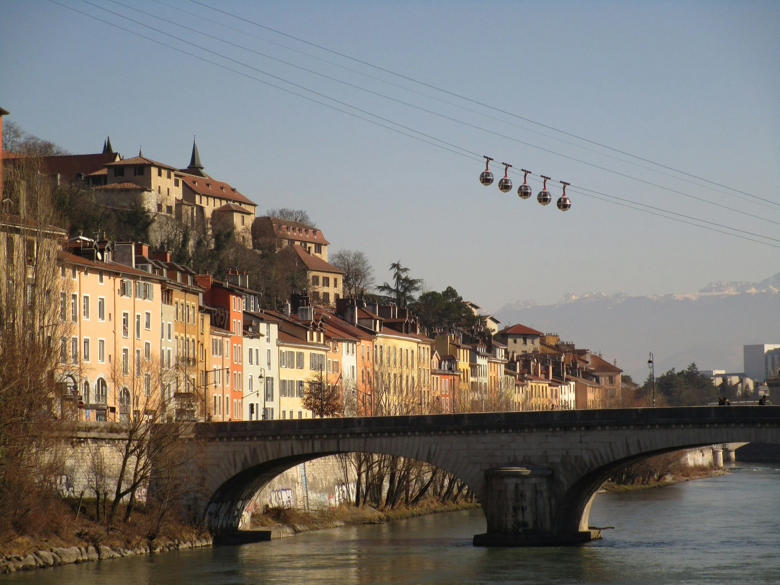 Un pont au-dessus d'une rivière, avec des bâtiments colorés le long de la rive et des cabines de téléphérique suspendues dans le ciel, avec des montagnes en arrière-plan.