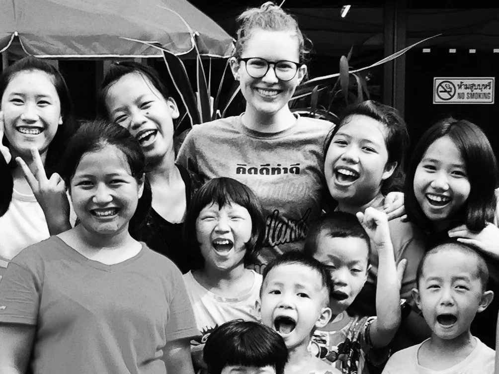 A group of children and a young woman, possibly a teacher or volunteer, smiling and laughing together outdoors. The children are diverse in appearance, and they seem happy and excited.