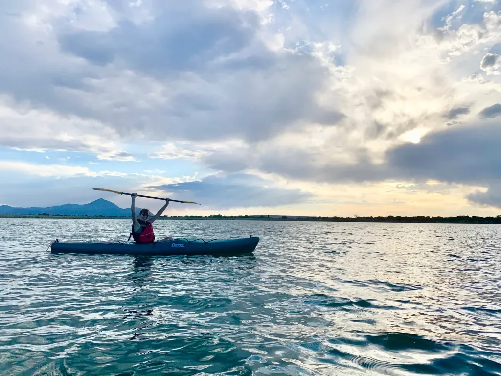 Person kayaking on a large body of water during sunset, holding a paddled above their head, with mountains and a cloudy sky in the background.