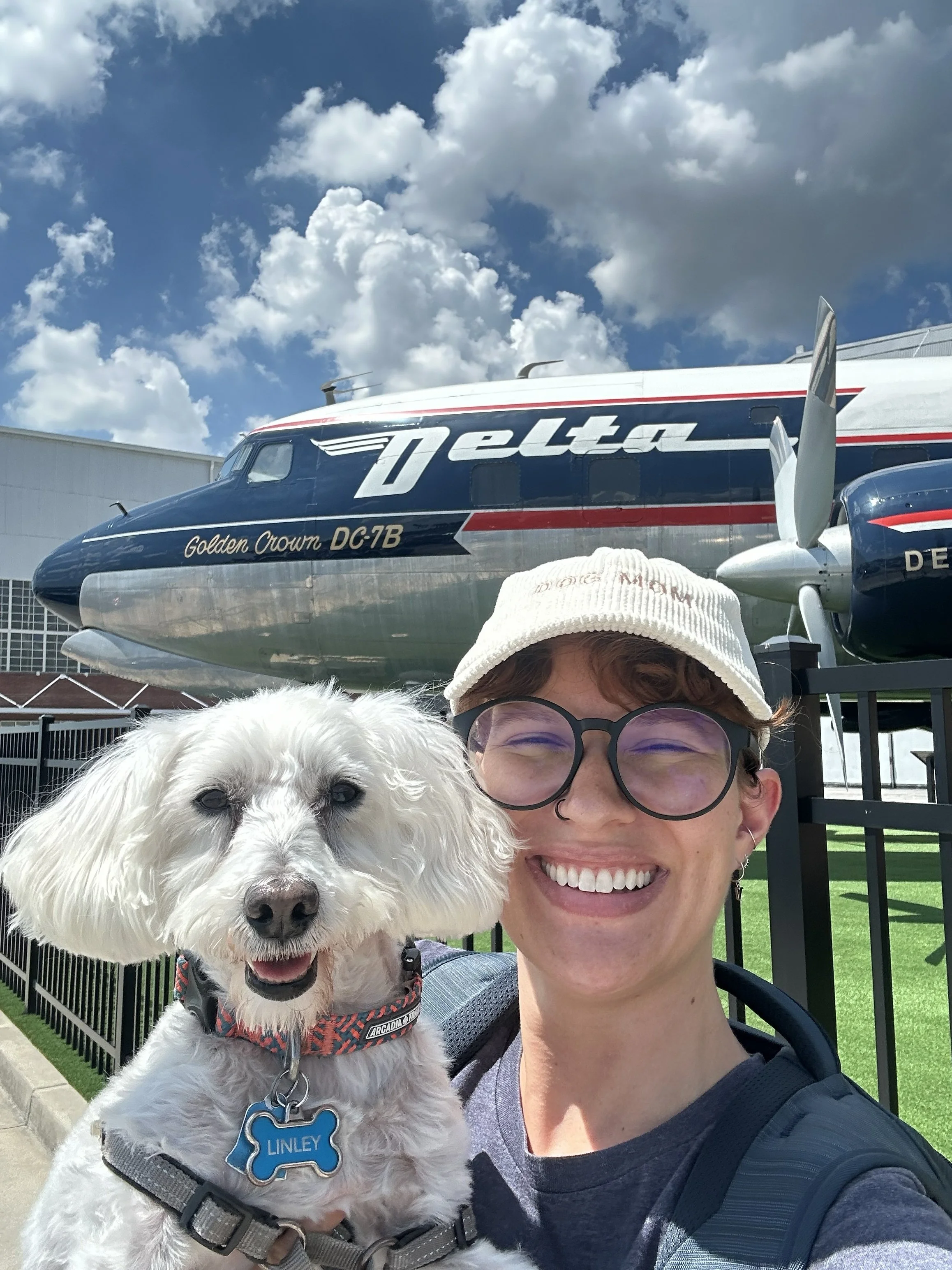A smiling woman with glasses, a beige cap, and a gray shirt taking a selfie with her white dog in front of a Delta airplane displayed as a static exhibit on a lawn, with cloudy skies above.