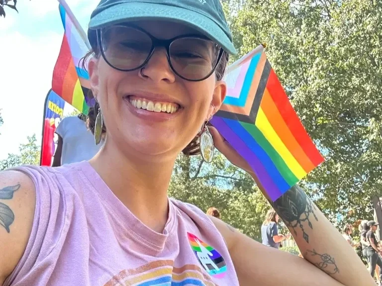 A person outdoors at a Pride event, smiling, wearing glasses, a cap, a pink sleeveless shirt, and rainbow-themed accessories including a rainbow flag and a rainbow-colored pin.