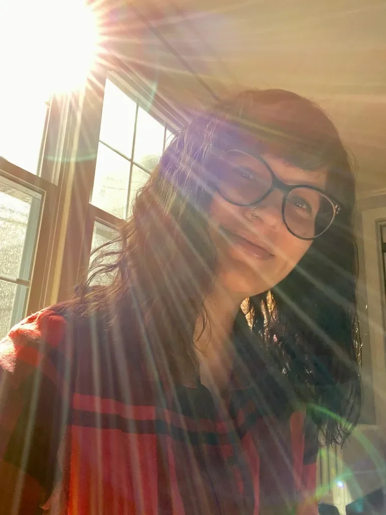 A woman with glasses and wavy dark hair taking a selfie in sunlight, near a window.