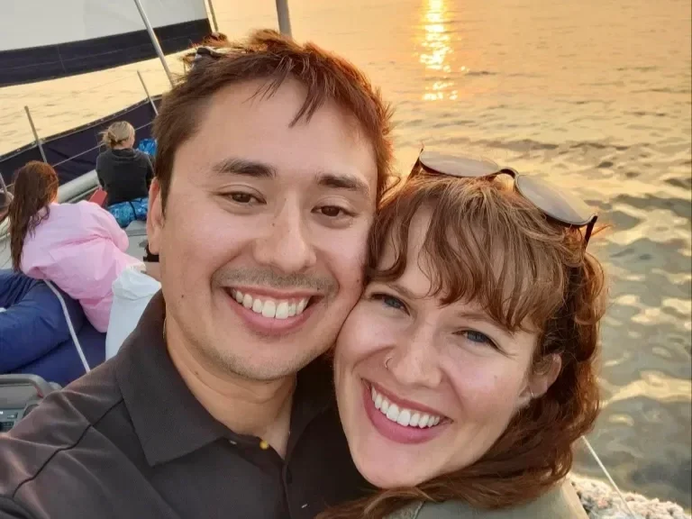 A smiling couple taking a selfie on a boat at sunset, with other passengers and the water behind them.