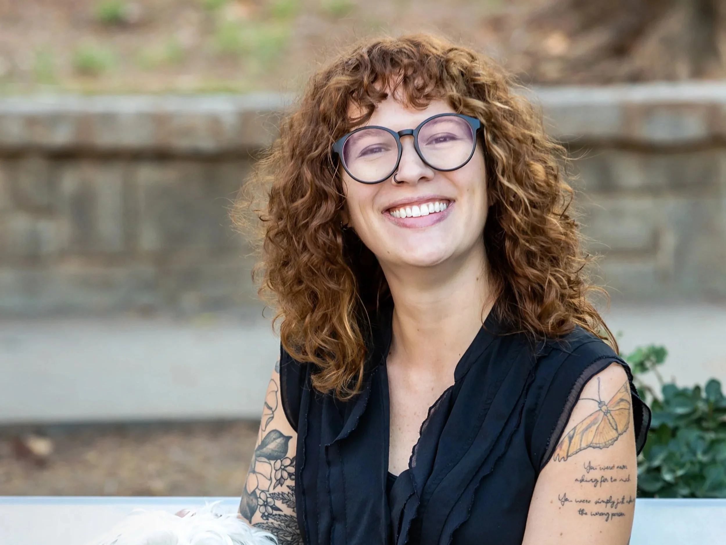 A woman with curly red hair, glasses, and tattoos on her arms, smiling outdoors.