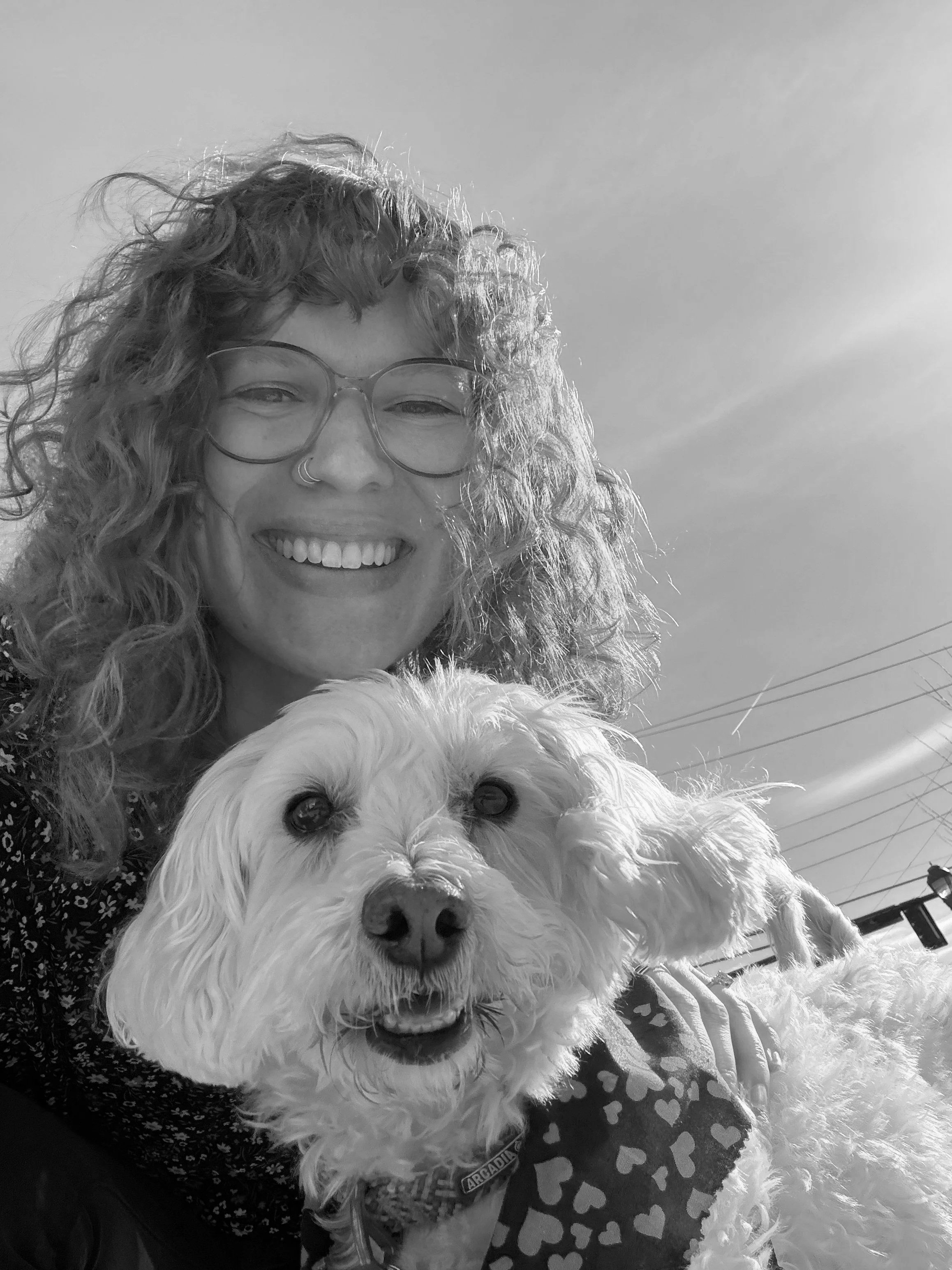 A woman with curly hair and glasses smiling next to a white fluffy dog with dark eyes and nose outdoors under a clear sky.