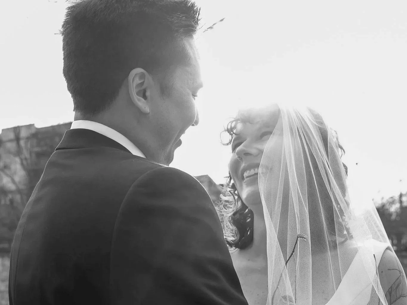 A black-and-white photo of a coupleon their wedding day, smiling at each other. The man is dressed in a suit, and the woman has curly hair with a veil covering part of her face. They are looking at each other with happiness, sunlight creating a bright glow around them.
