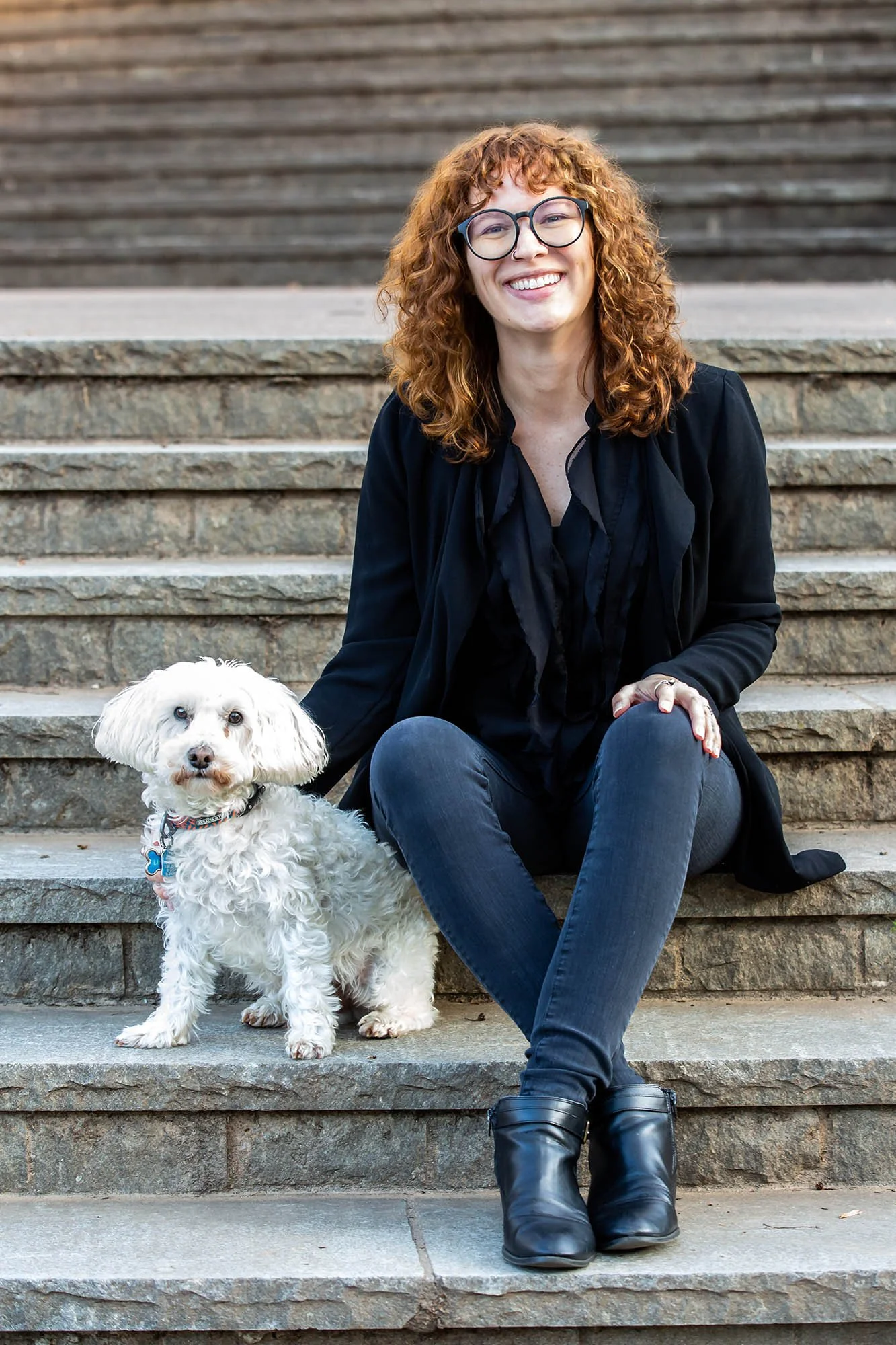 A woman with curly red hair, glasses, and a nose ring sitting on outdoor stone stairs with a white fluffy dog.