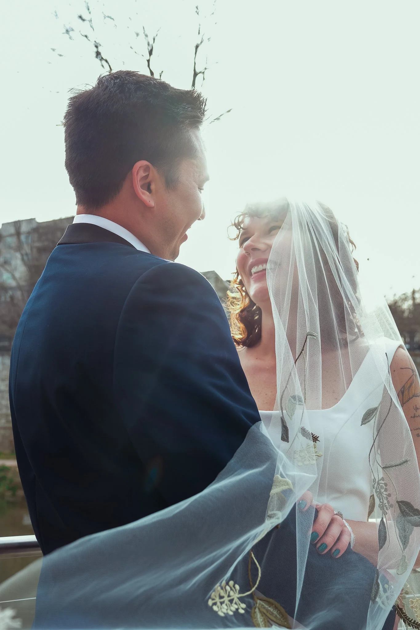 A couple on their wedding day smiling at each other, with a woman wearing a wedding dress and veil and a man in a dark suit outdoors with trees in the background.