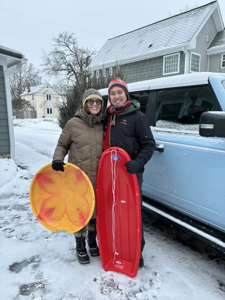 Two people dressed in winter clothing standing outside in snow, smiling while holding a yellow sled and a red sled in front of a gray house and a blue vehicle.