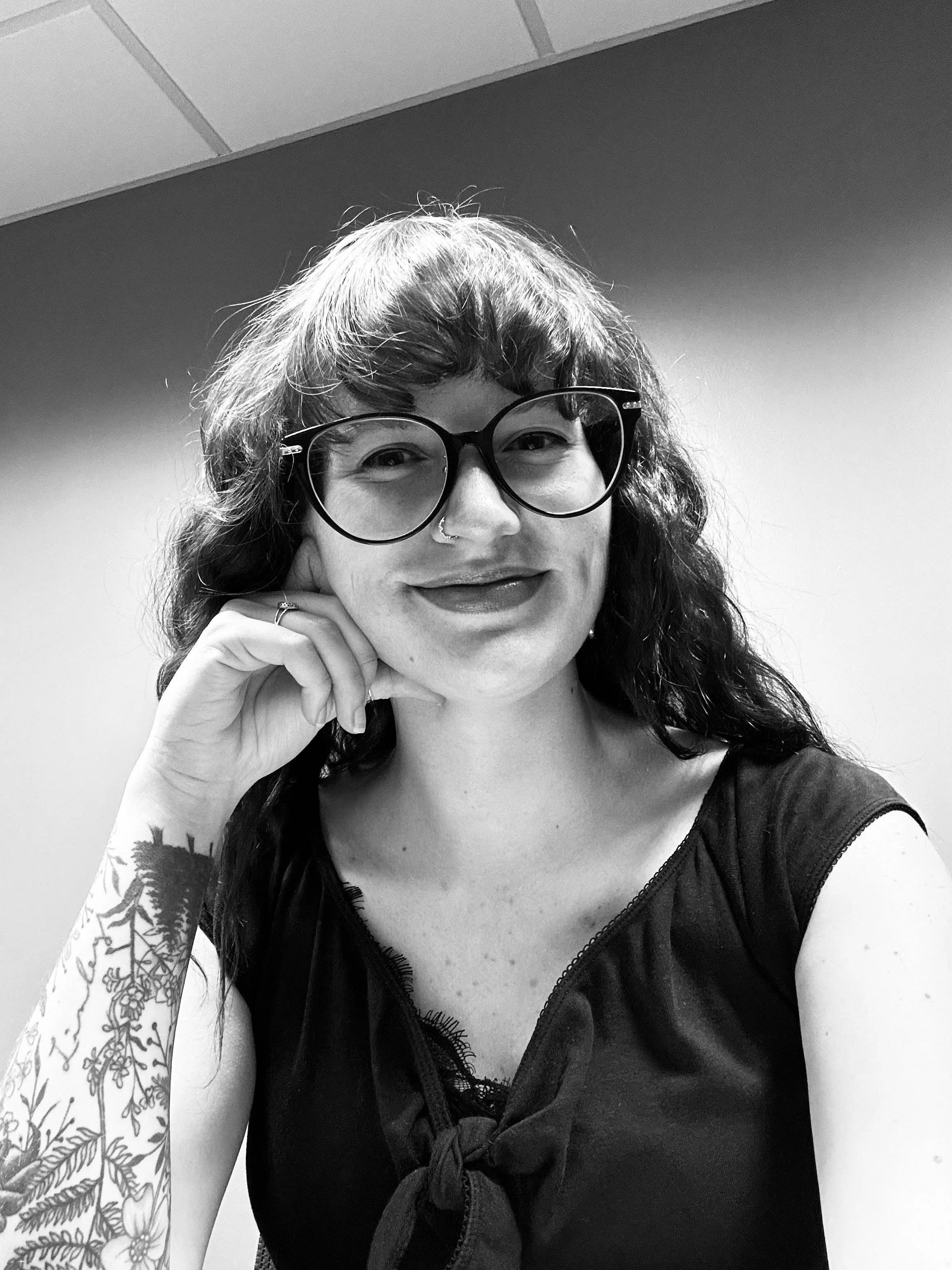 A woman with curly hair and glasses smiling, resting her chin on her hand, wearing a dark top, a nose ring, and a sleeve tattoo of flowers and leaves.