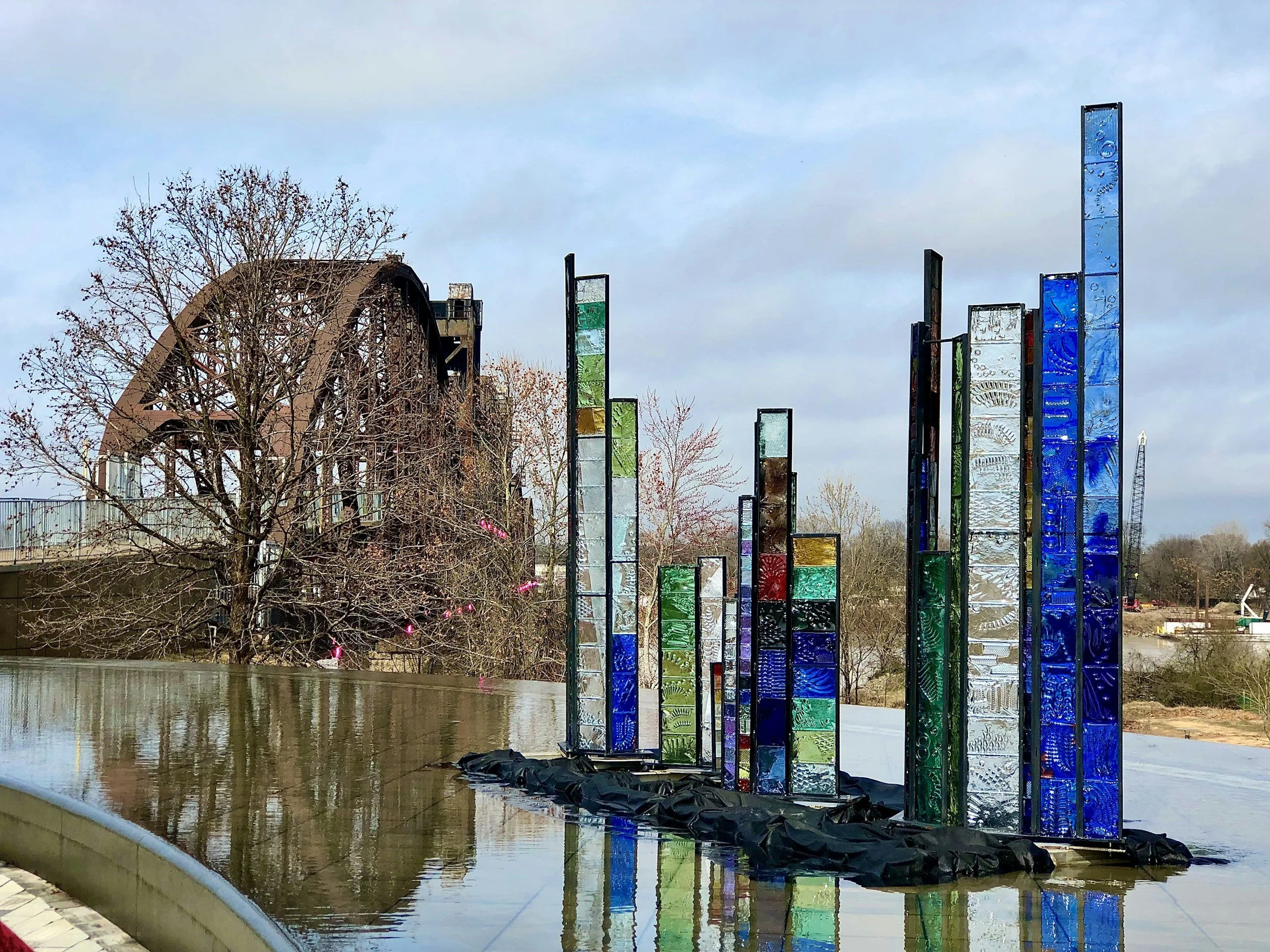 E. A. Lusinsaano Colorful glass sculpture installation with tall rectangular panels near a reflective water feature, with a brown arched building and trees in the background.