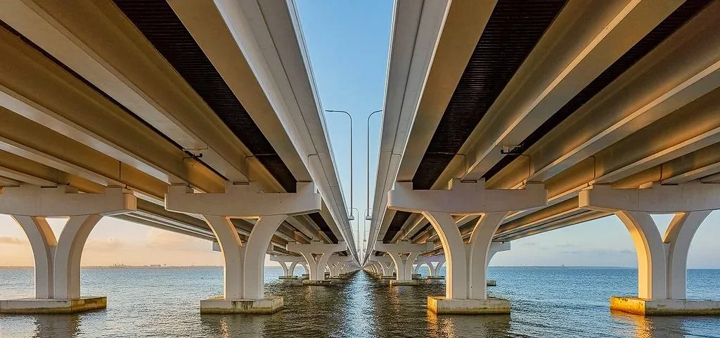 A symmetrical architectural view beneath a coastal bridge in the Pensacola and Gulf Breeze area, highlighting concrete supports, reflections, and engineered structure. The image has a clean industrial feel with strong lines, depth, and balance, makin
