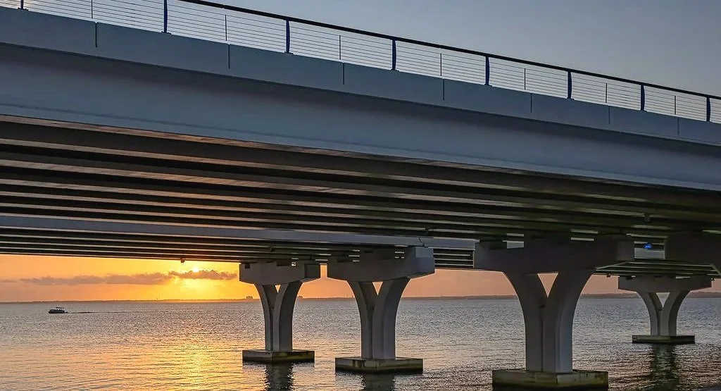 A moody late evening bridge scene from the Gulf Breeze Bridge, featuring dark structural lines against a brightening sky. The contrast between shadow, water, and sunrise color gives this photograph a strong industrial coastal atmosphere. This piece w