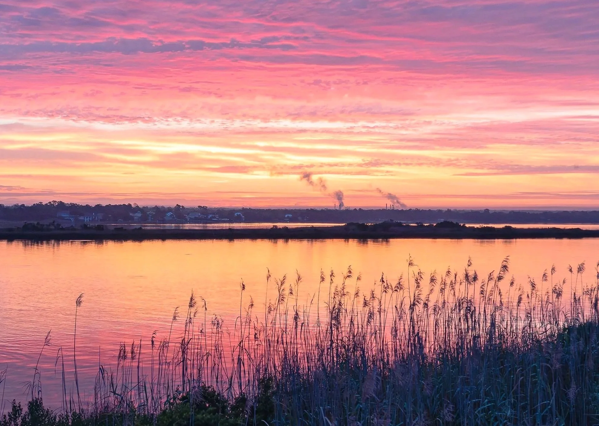 A breathtaking blue hour sunrise captured from the Milton-Pensacola Bridge, Florida. Vivid bands of magenta, coral pink, and golden orange stretch across the sky and reflect perfectly in the glassy surface of Pensacola Bay. Silhouetted marsh grass an