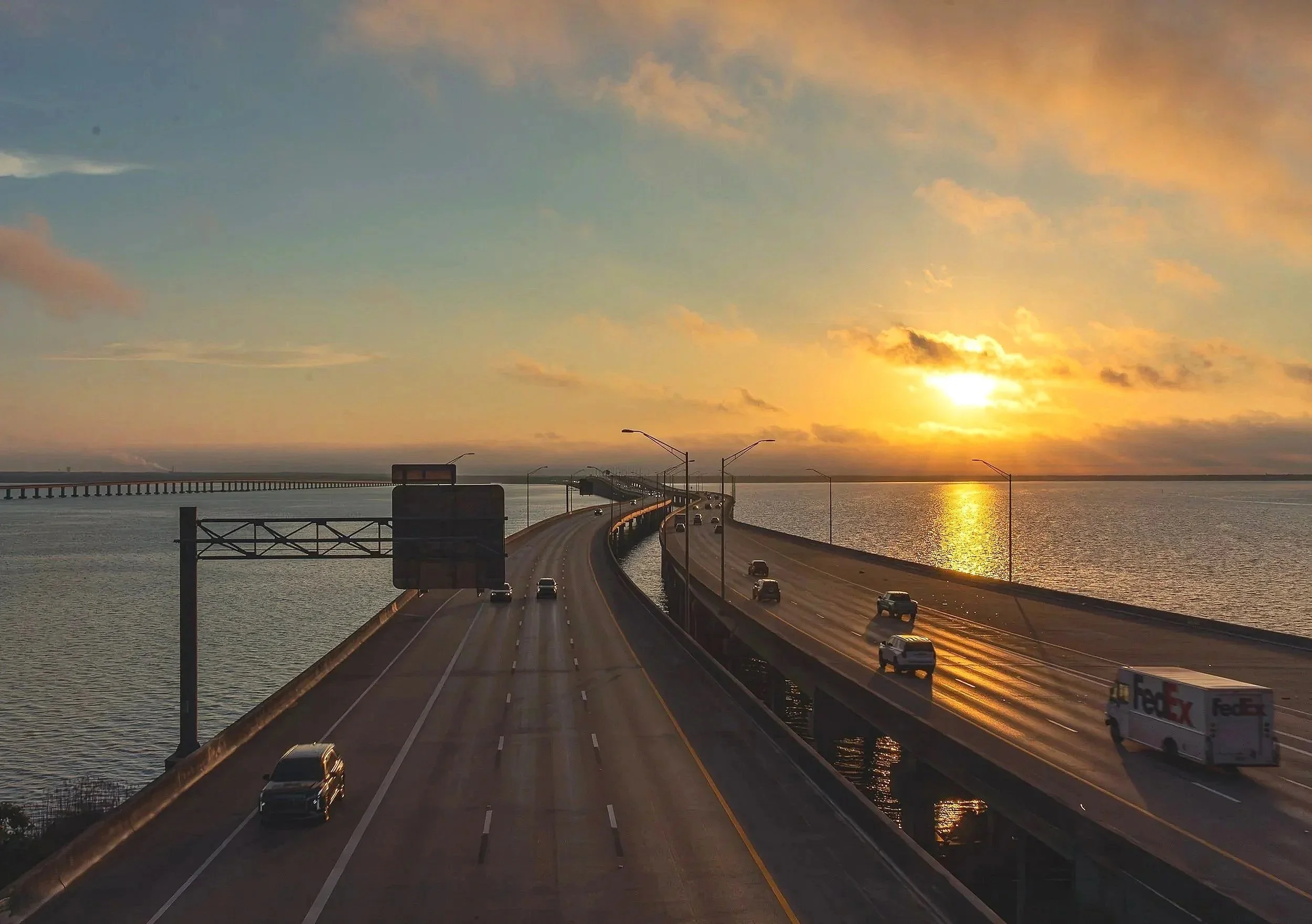 A dramatic Gulf Coast sunrise captured over the Pensacola Bay Bridge, with golden morning light reflecting across the water and roadway. This image blends coastal landscape photography with strong architectural lines, creating a peaceful yet powerful