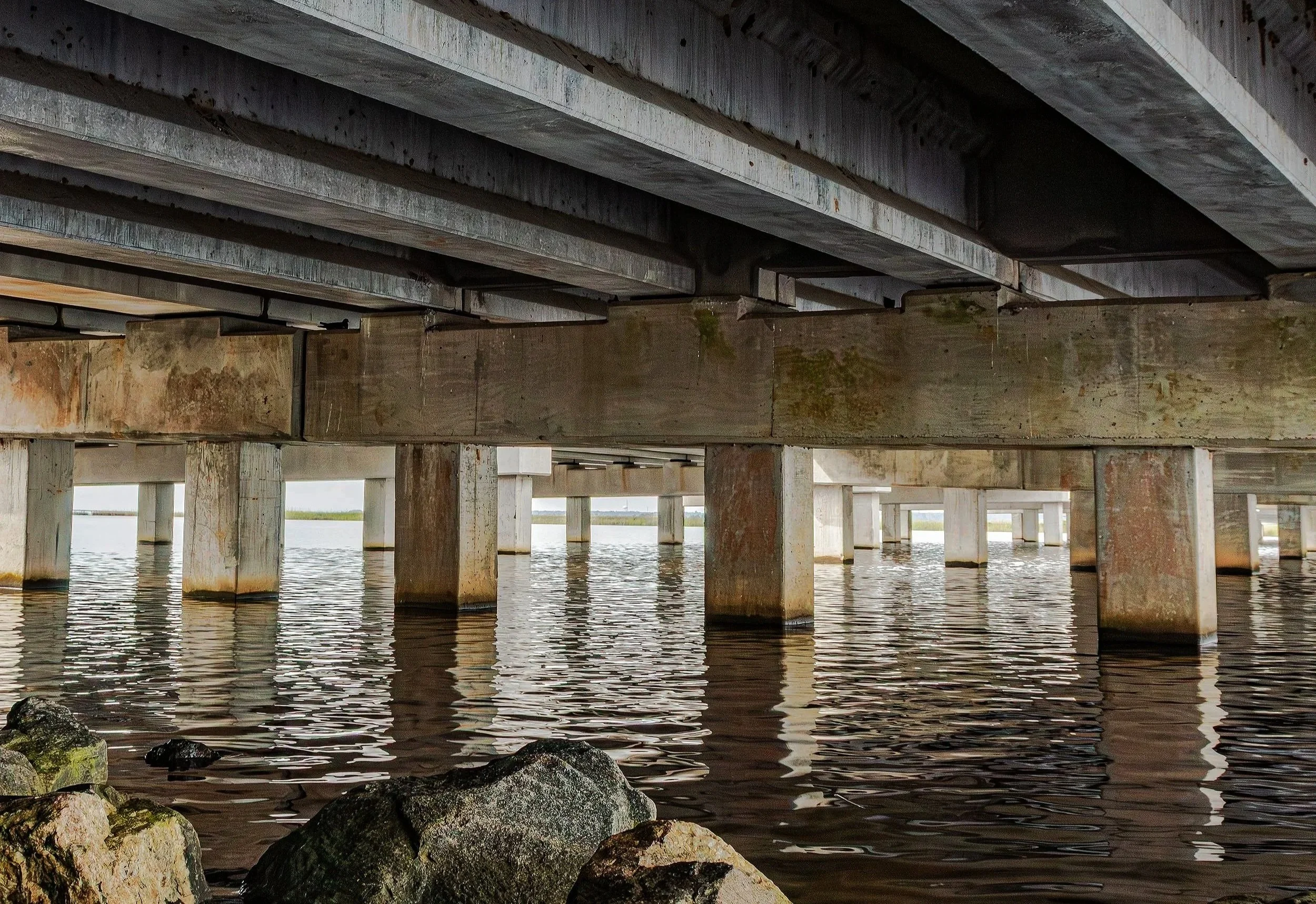 The geometric underside of a Pensacola-area bridge frames a colonnade of concrete pillars receding into the distance, mirrored in the still water below. Rock formations anchor the foreground as morning light filters through the structure. Gulf Coast 
