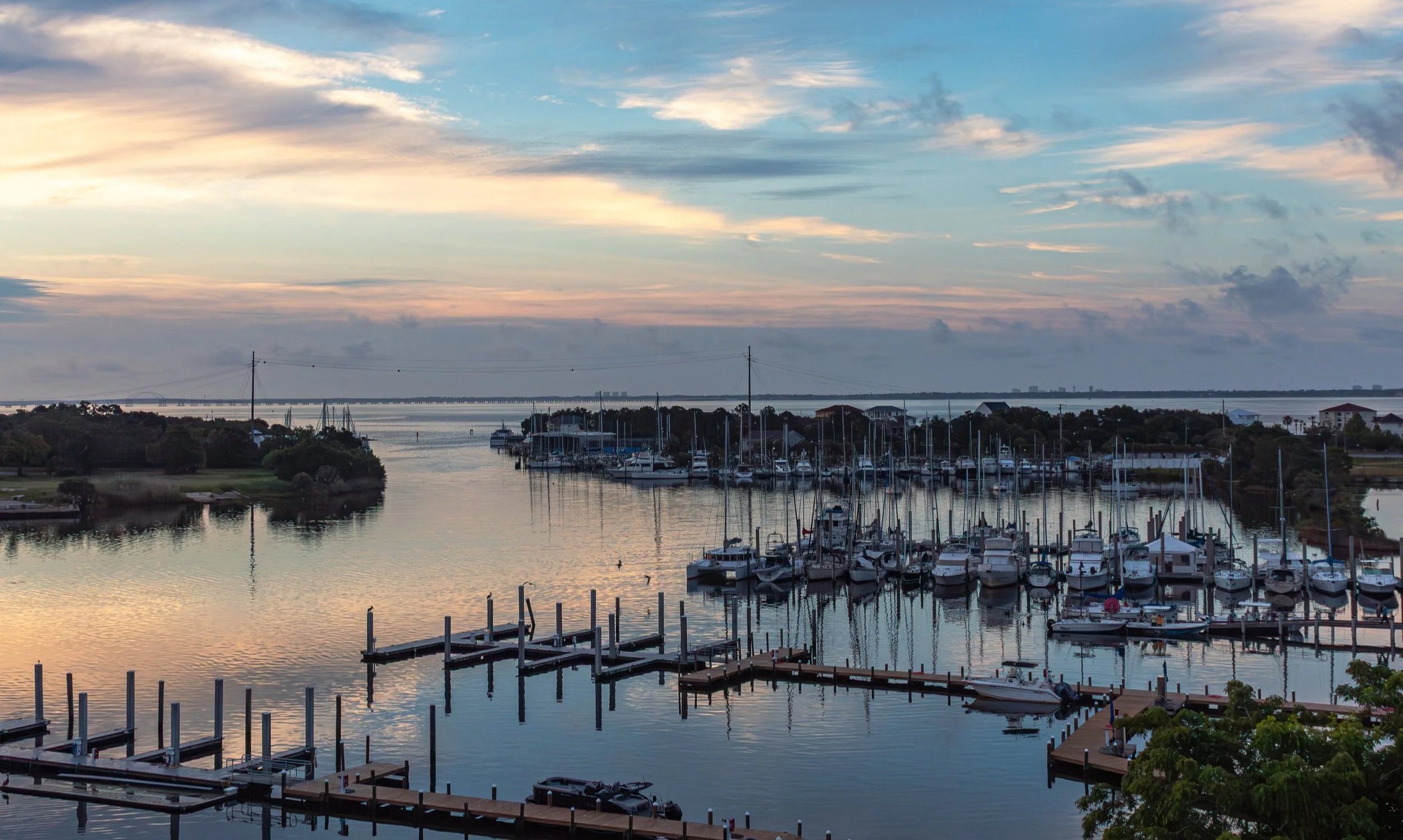 The full breadth of Pensacola Bay opens from the Barrancas Avenue bridge on a luminous early morning — sailboat masts line the right arc of the marina, empty dock pilings extend into the calm foreground water, and a layered pink, lavender, and blue s