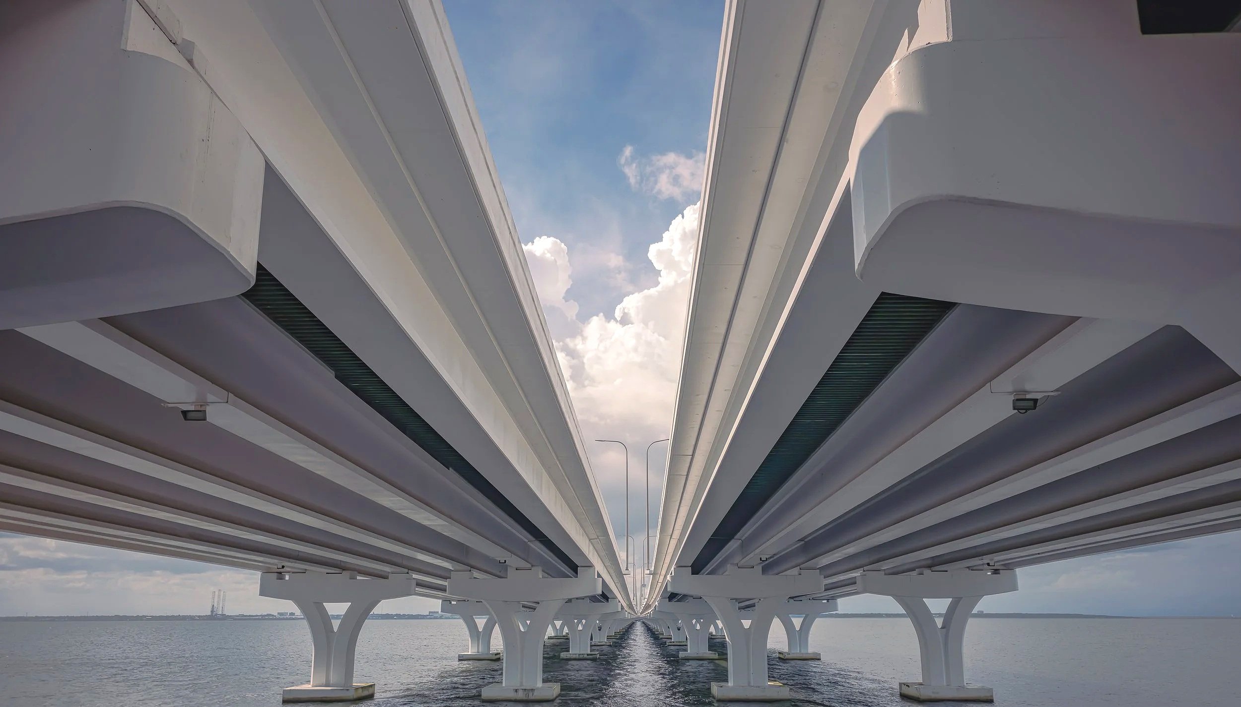 A symmetrical architectural view beneath a coastal bridge in the Pensacola and Gulf Breeze area, highlighting concrete supports, reflections, and engineered structure. The image has a clean industrial feel with strong lines, depth, and balance, makin
