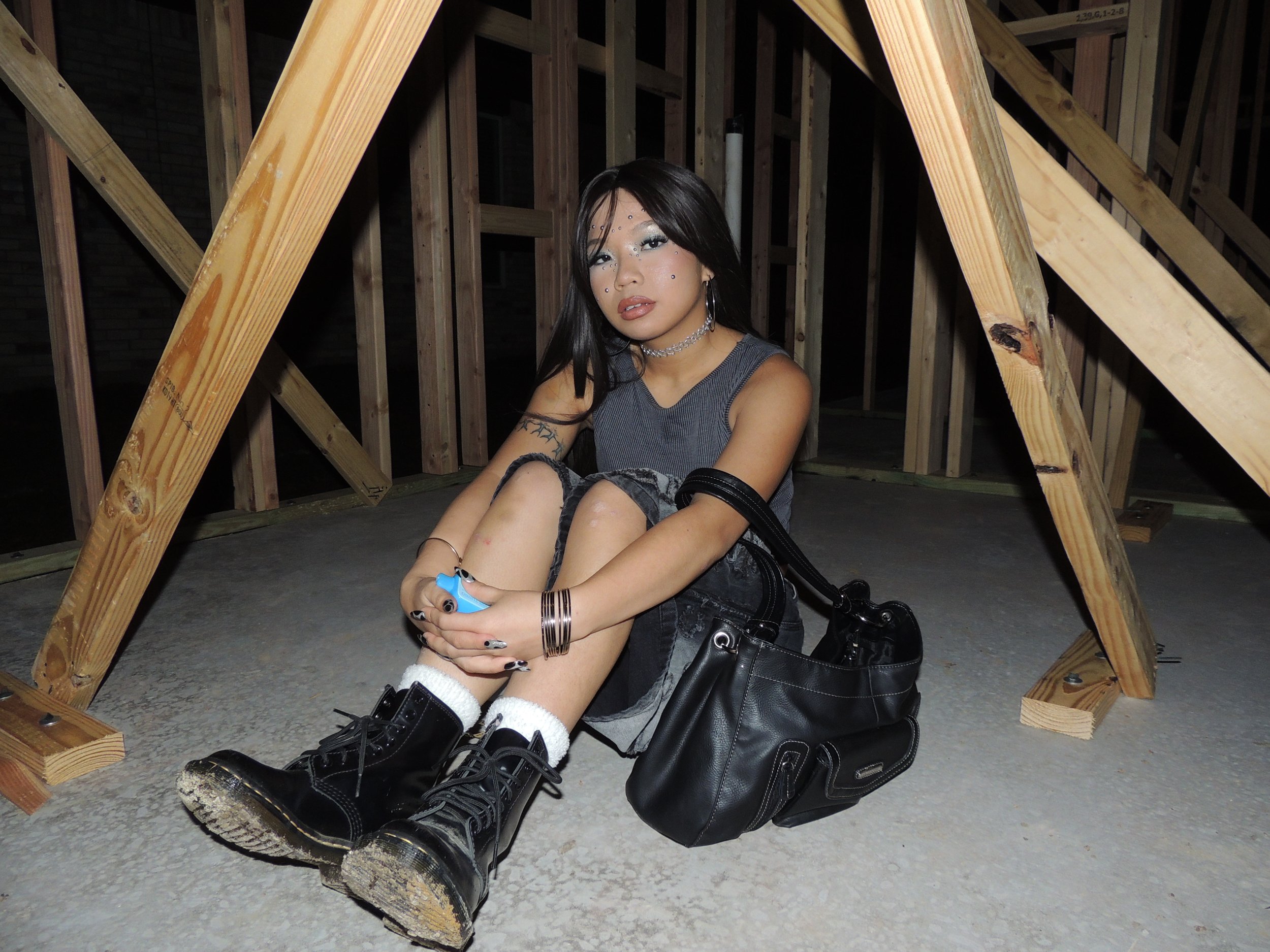 A young woman with dark hair and makeup sits on the concrete floor of a wooden attic, surrounded by exposed wooden framing. She wears a sleeveless gray top, black shorts, black combat boots, accessories including earrings, a choker, bracelets, and ma