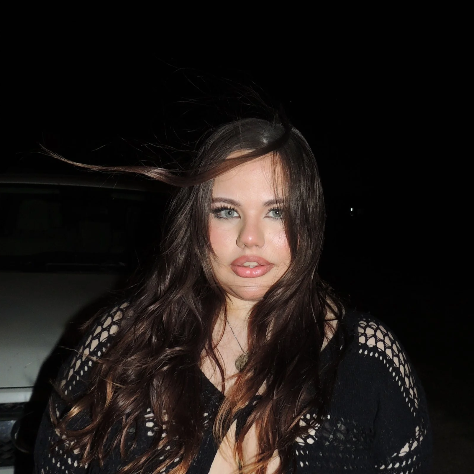 A woman with long wavy brown hair and blue eyes poses outdoors at night with a dark background and a blurred vehicle behind her, wearing a black lace top.