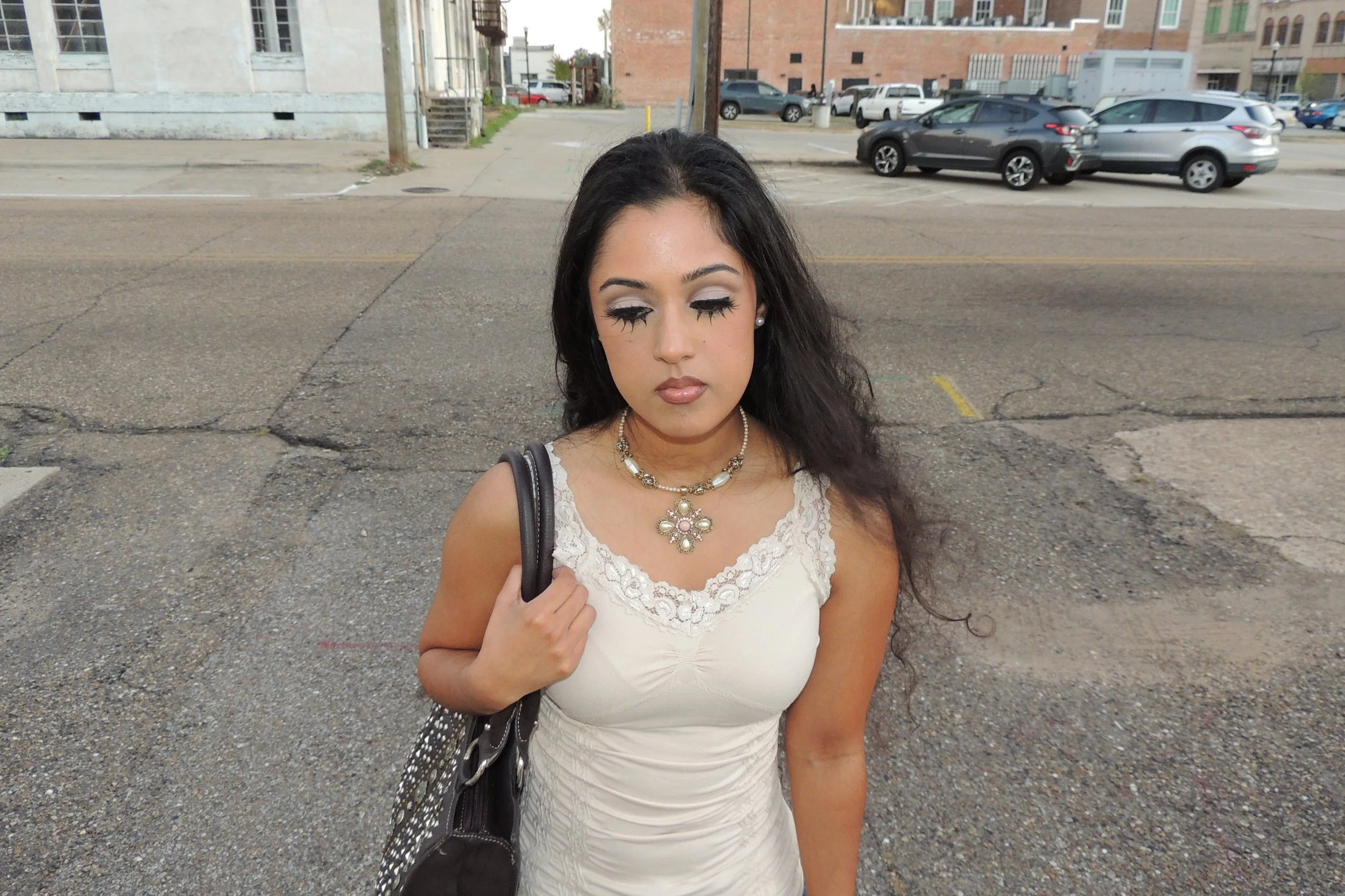 A woman with long dark hair, dressed in a white lace top, wearing pearl jewelry and carrying a black shoulder bag, standing on an urban street with parked cars and buildings in the background, eyes closed.