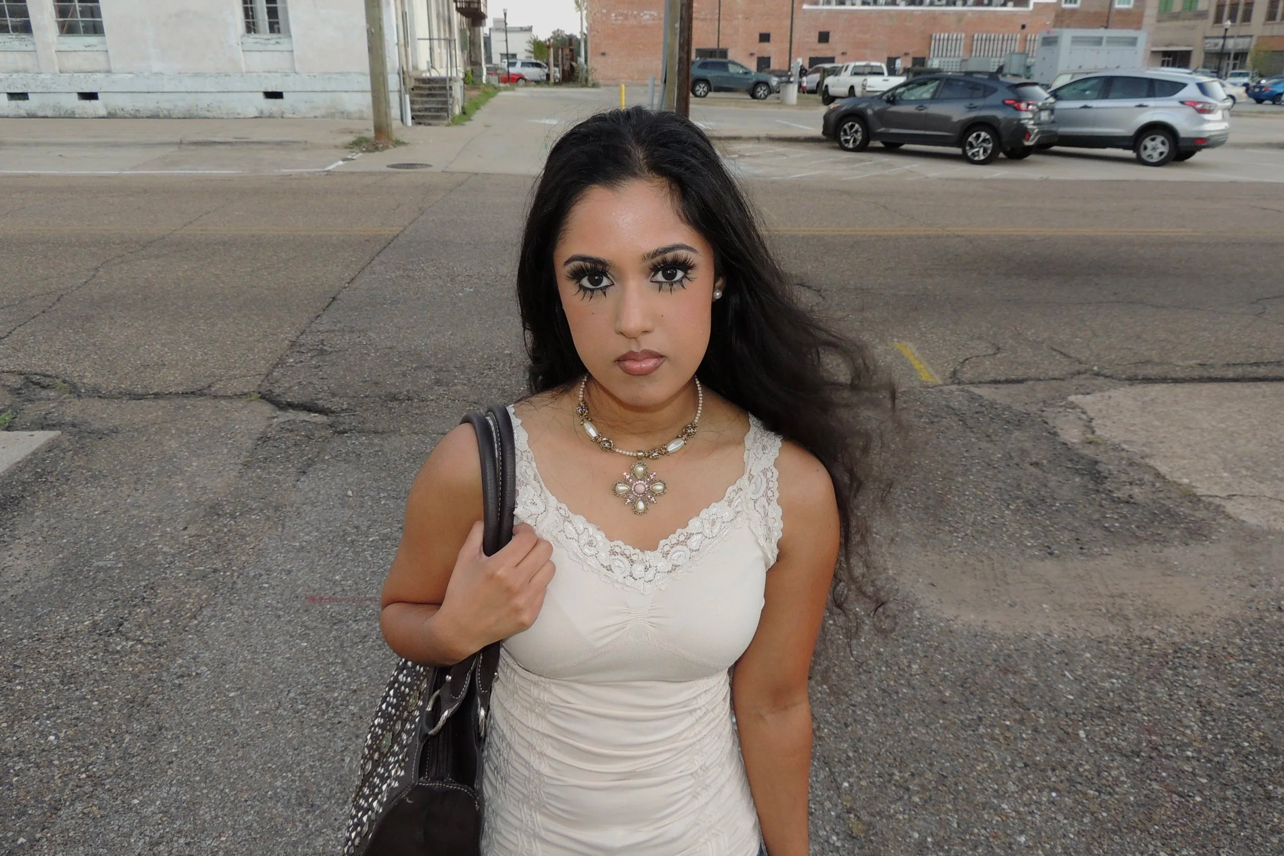 A woman with long black hair and makeup, wearing a white lace dress and jewelry, stands outdoors on a street with parked cars and buildings in the background.