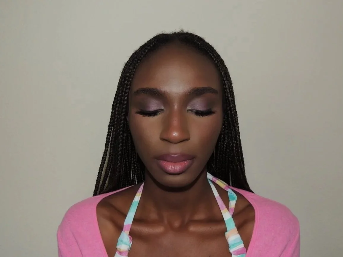 Young woman with braided hair, closed eyes, light makeup, wearing pink top with colorful striped straps against a plain wall.