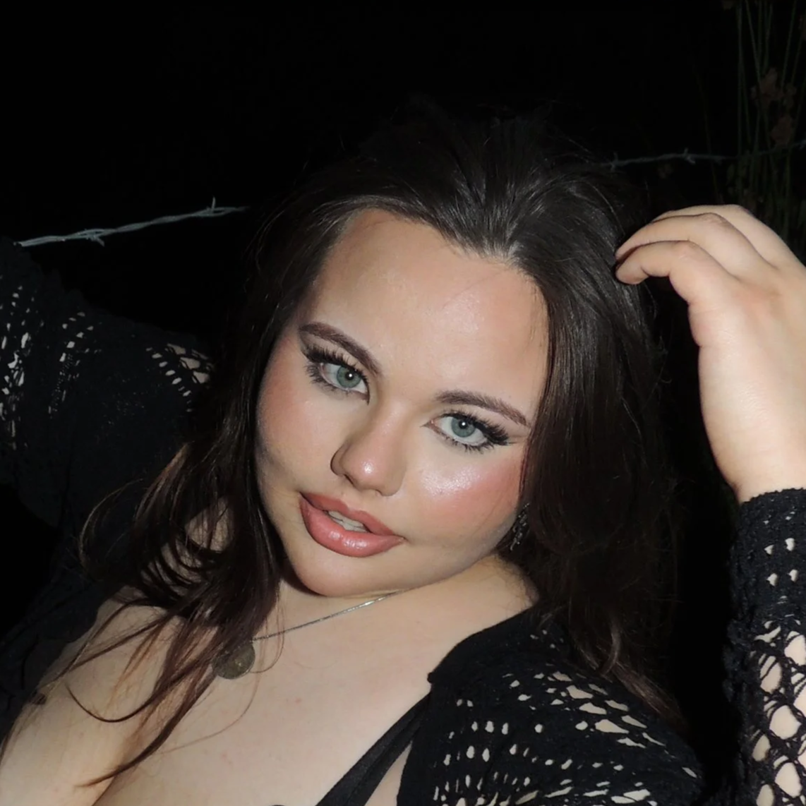 Close-up photo of a woman with long, dark hair and blue eyes, wearing makeup, a black lace top, and a necklace, posing outdoors at night.