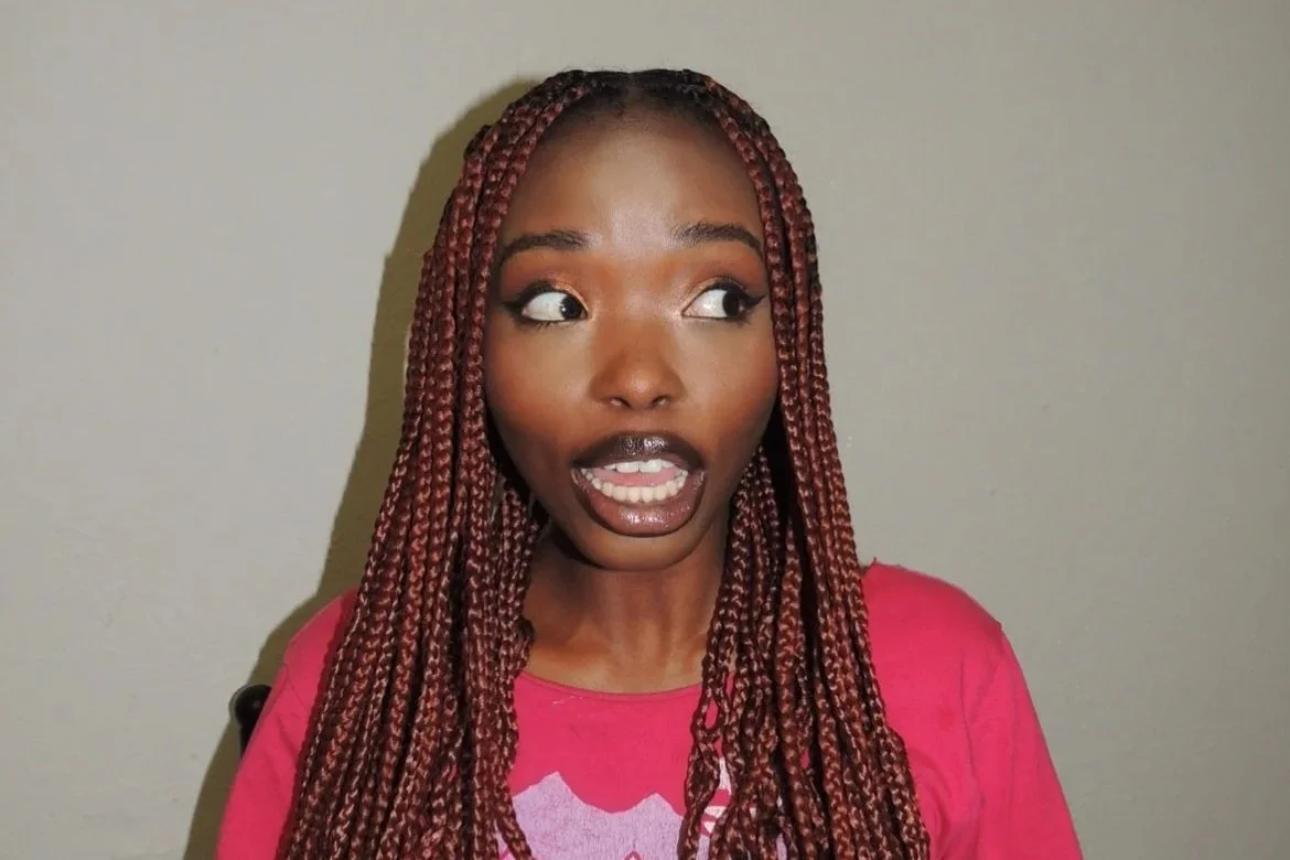 A woman with long, reddish-brown box braids wearing a pink shirt, making a surprised or playful face, against a plain background.