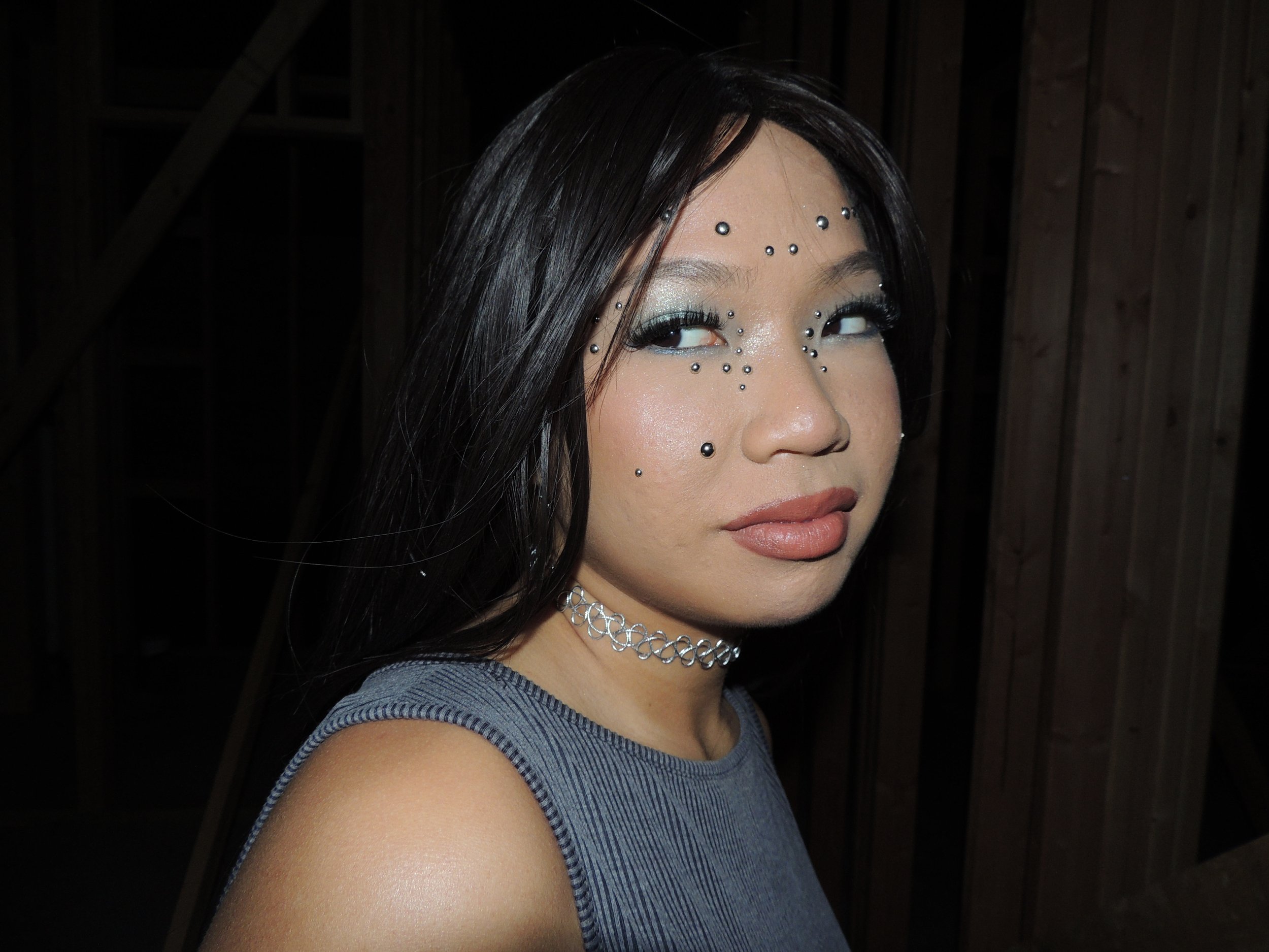 A woman with dark hair and makeup, adorned with rhinestones on her face, wearing a choker and a sleeveless top, indoors with wooden panels in the background.