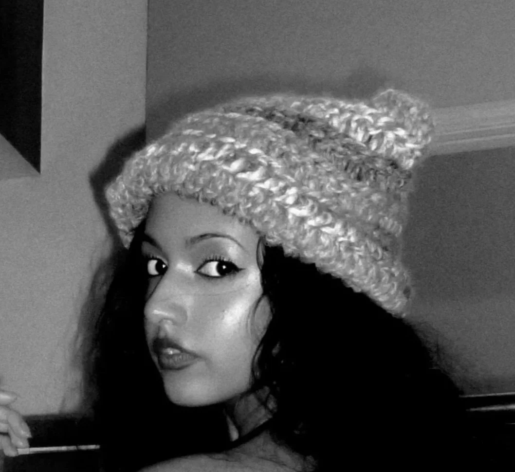 A woman with dark wavy hair wearing a thick, knitted hat with a pom-pom on top, looking over her shoulder in a black and white photo.