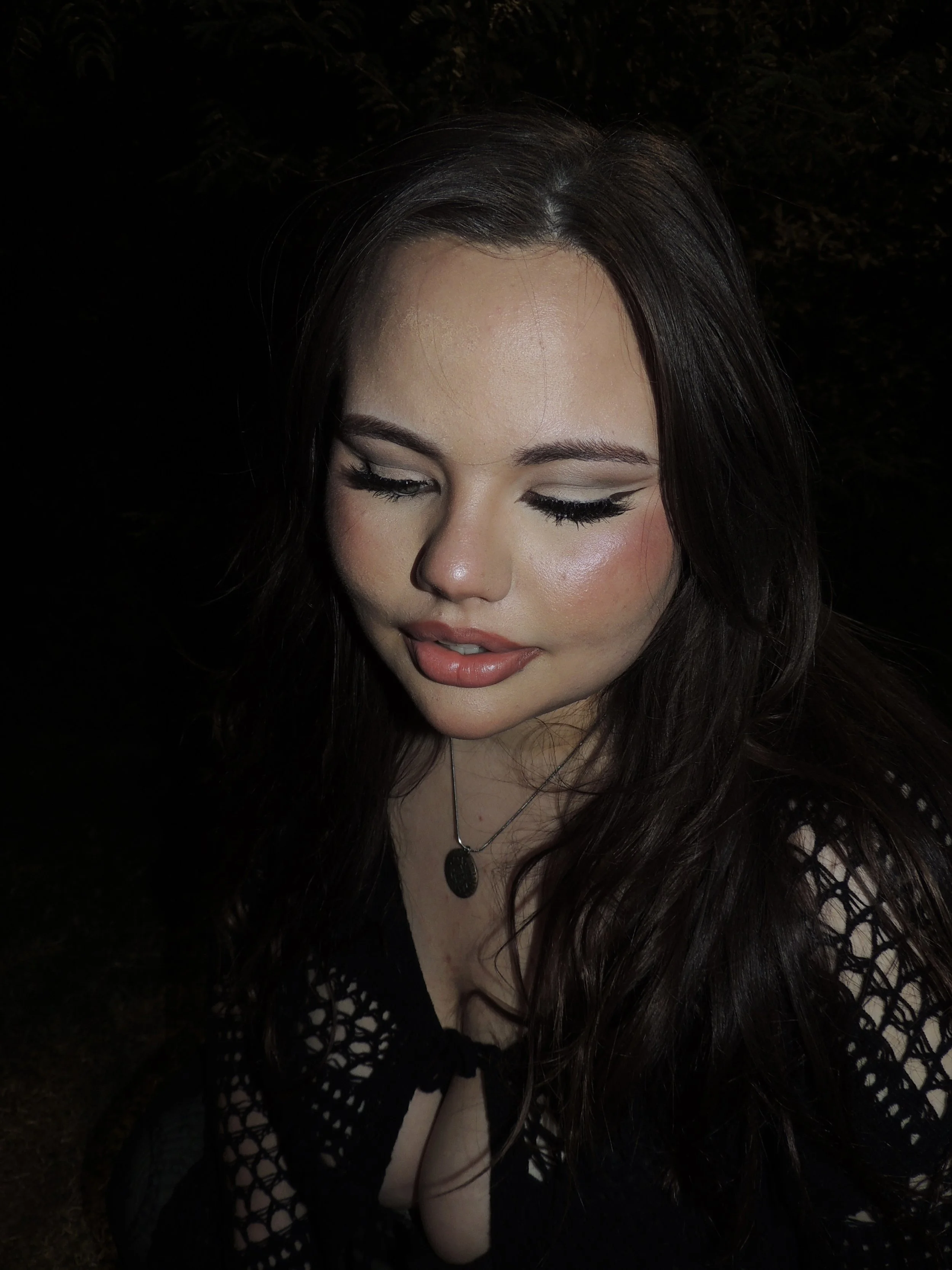 A young woman with long dark hair, closed eyes, and makeup, wearing a black crochet top and a necklace, looking down.
