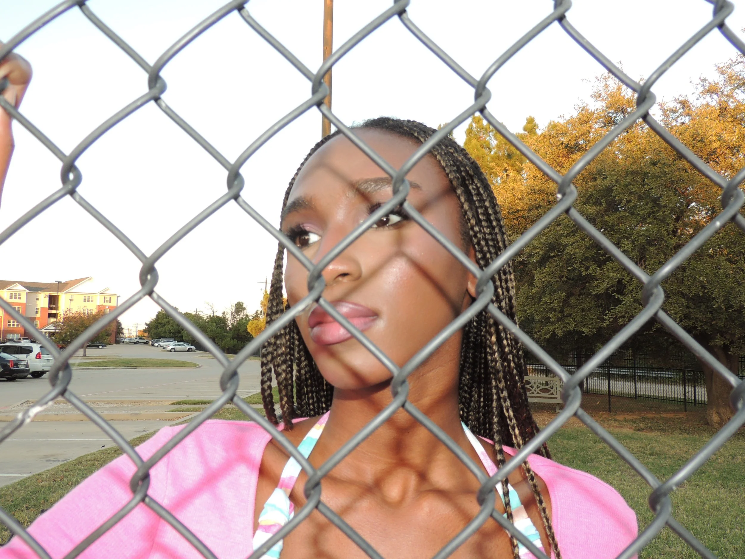A woman with braided hair wearing a pink top and colorful swimsuit, standing behind a chain-link fence in an outdoor setting with trees and parked cars in the background.