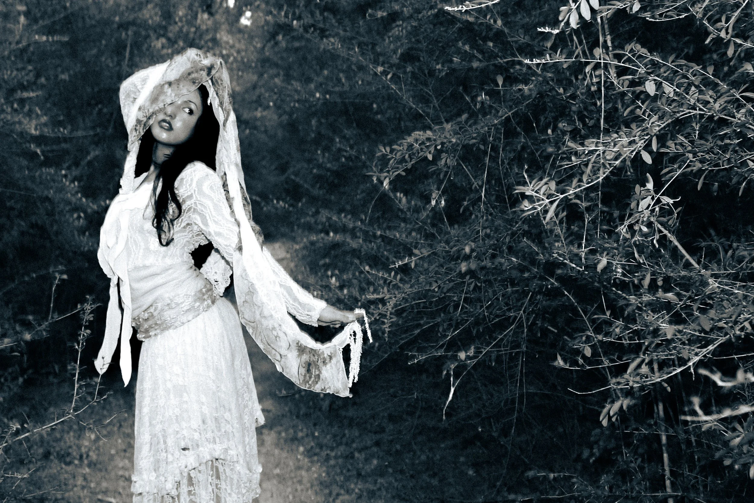 A woman in a white lace dress and a large hat standing in a wooded area.