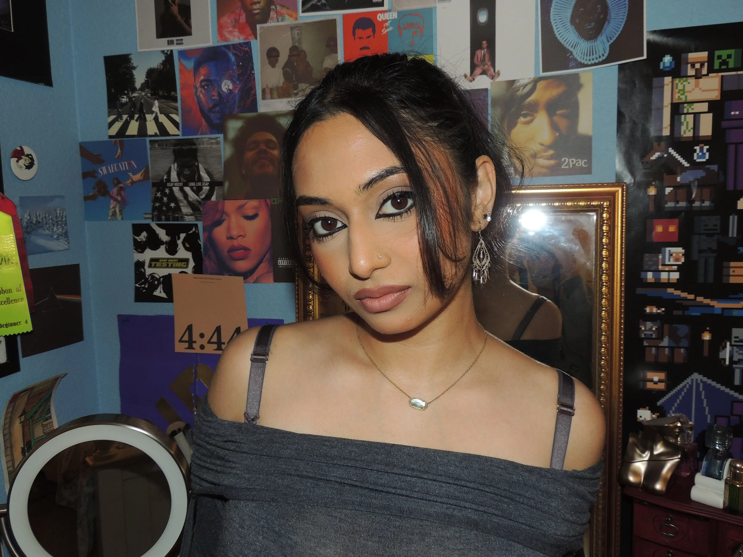 A young woman with dark hair and makeup posing indoors against a wall with various posters, including a queen and a 2Pac poster, and a framed mirror behind her.