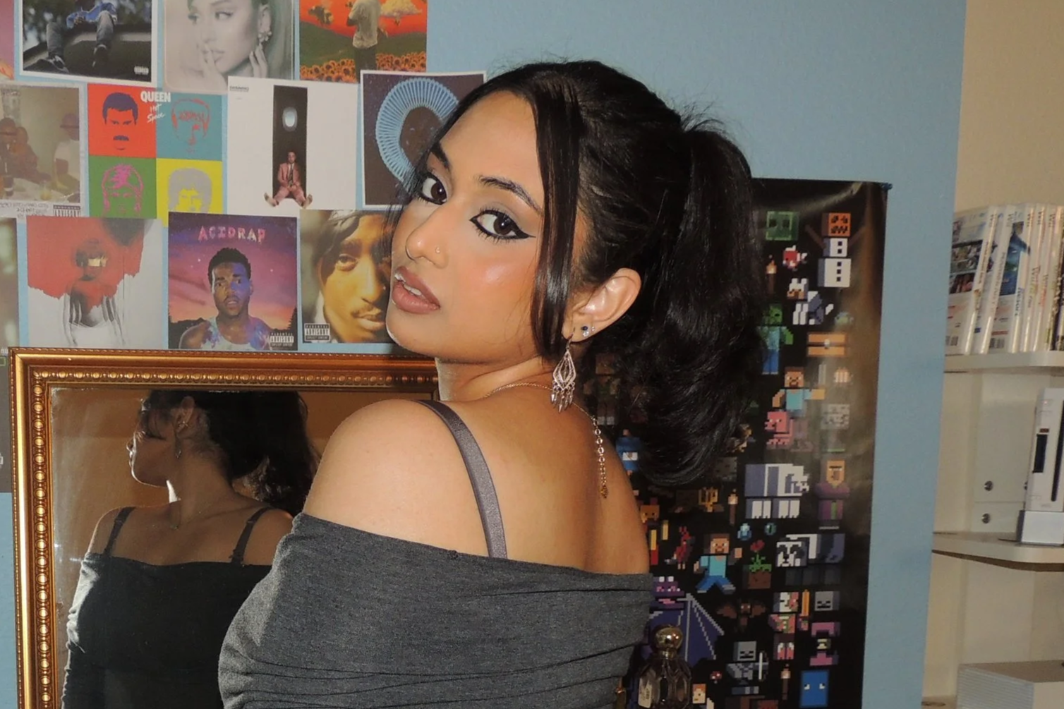 A young woman with dark hair in a ponytail is looking over her shoulder at the camera. She has dramatic eye makeup, a nose piercing, and is wearing earrings, a necklace, and a gray off-shoulder top. The background includes a mirror, a colorful collage of album covers and posters, and shelves with books and boxes.