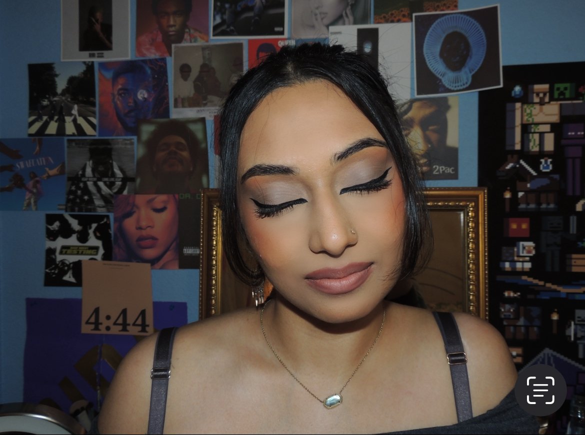 A woman with closed eyes and makeup, wearing a necklace, in front of a wall covered with posters and photos.
