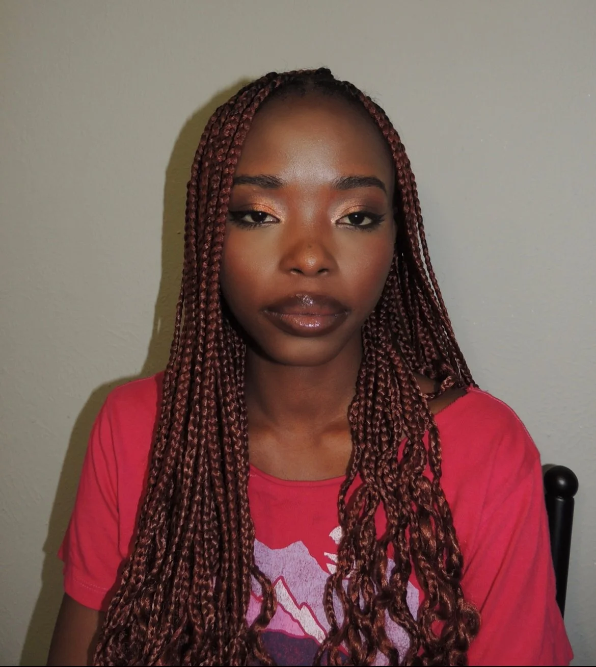 Close-up of a young woman with long reddish-brown braids, wearing makeup and a red T-shirt, against a plain wall background.