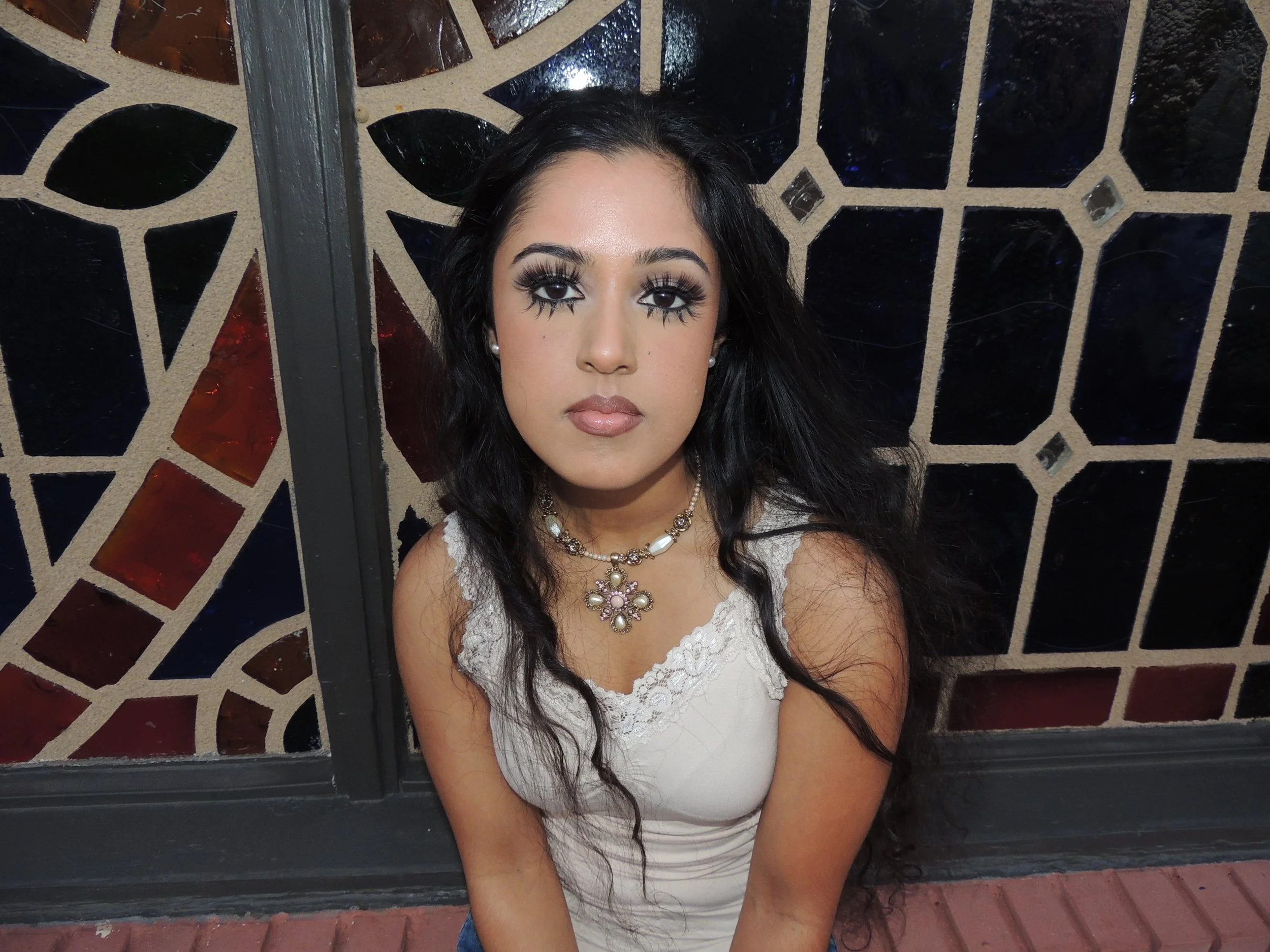 A young woman with long black hair and makeup featuring dark eyeliner and false eyelashes, wearing a white lace top, a necklace, and earrings, sitting in front of a stained glass window with red, black, and beige patterns.