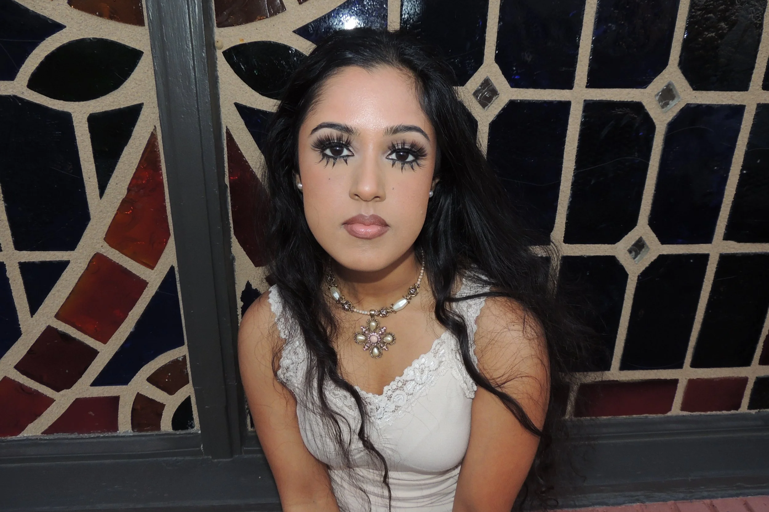 A woman with long dark hair and dramatic makeup, including bold eye makeup and lashes, wearing a white lace top, and a pearl and floral necklace, sitting in front of a decorative glass window with geometric and stained glass design.